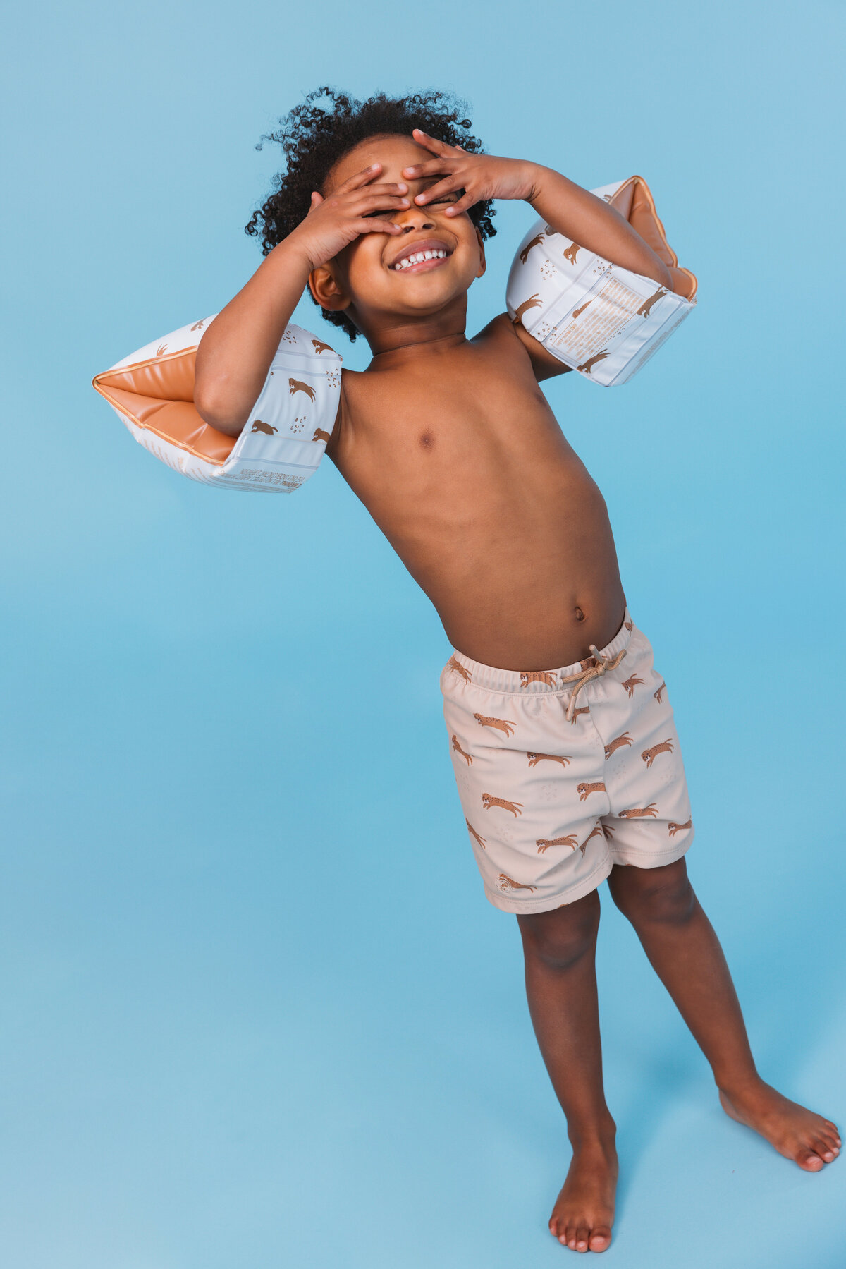 Smiling child wearing Swim Essentials beige floaties and matching swim shorts with panther print, posing playfully against a light blue background.