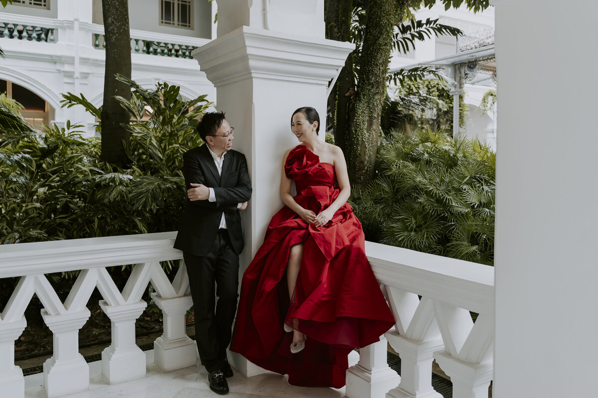 a woman in a red dress is sitting on the white railing while looking at a man in a black suit
