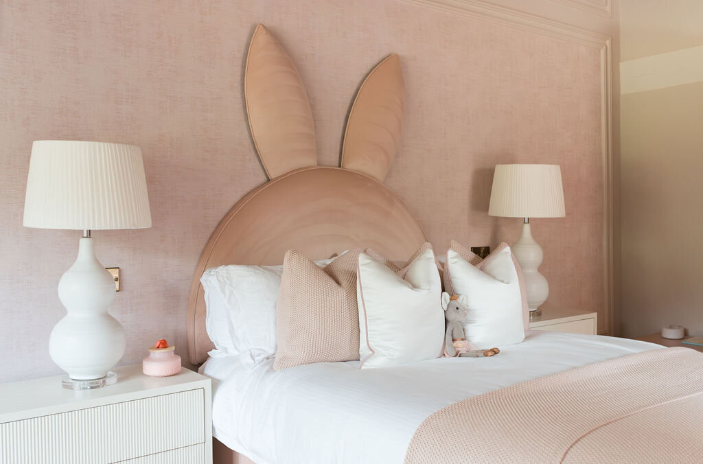 A cozy girl’s bedroom features a soft pink color palette, with a bed adorned with a unique headboard shaped like bunny ears. Two modern white nightstands with large white lamps flank the bed.