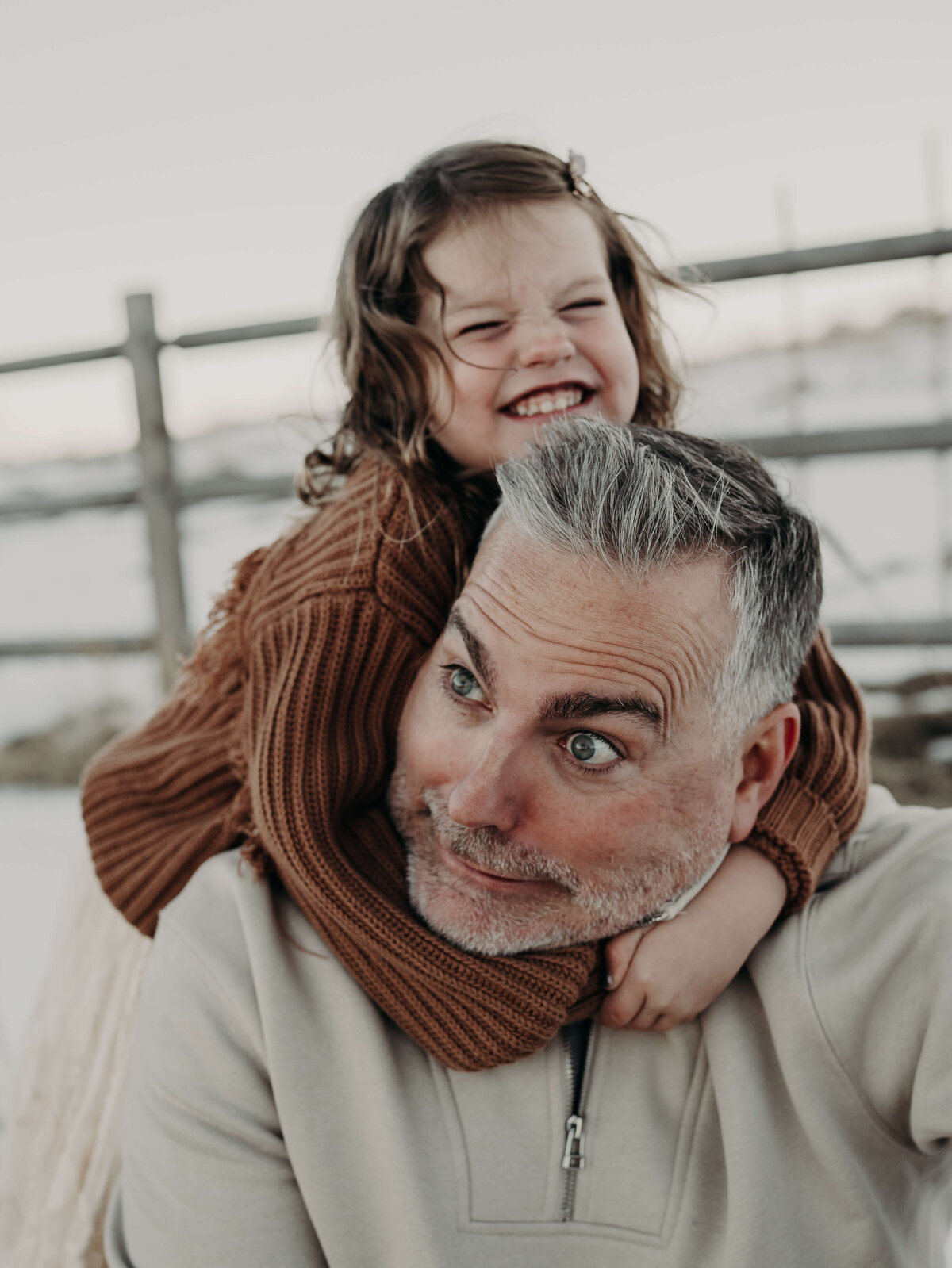 A sassy toddler jumps on her dad's back while wrapping her arms around his neck during a family photography session with snow and a farm fence in the background.
