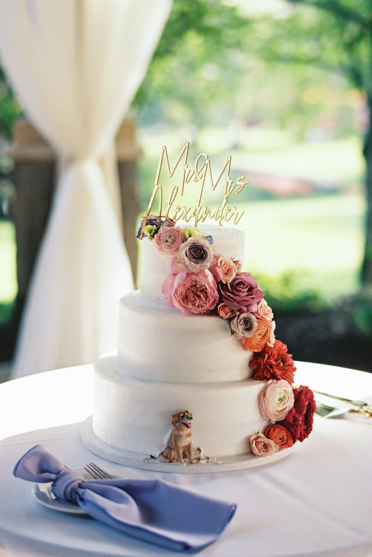 Three-tier wedding cake with cascading colorful florals and "Mr & Mrs Alexander" topper at Gervasi Vineyard.