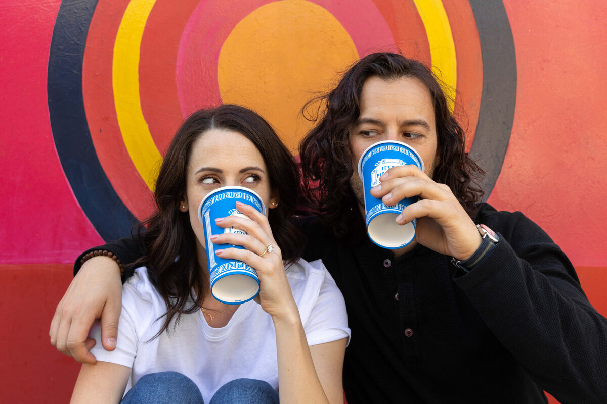 A person with their arm around their partner as they both drink from cups.