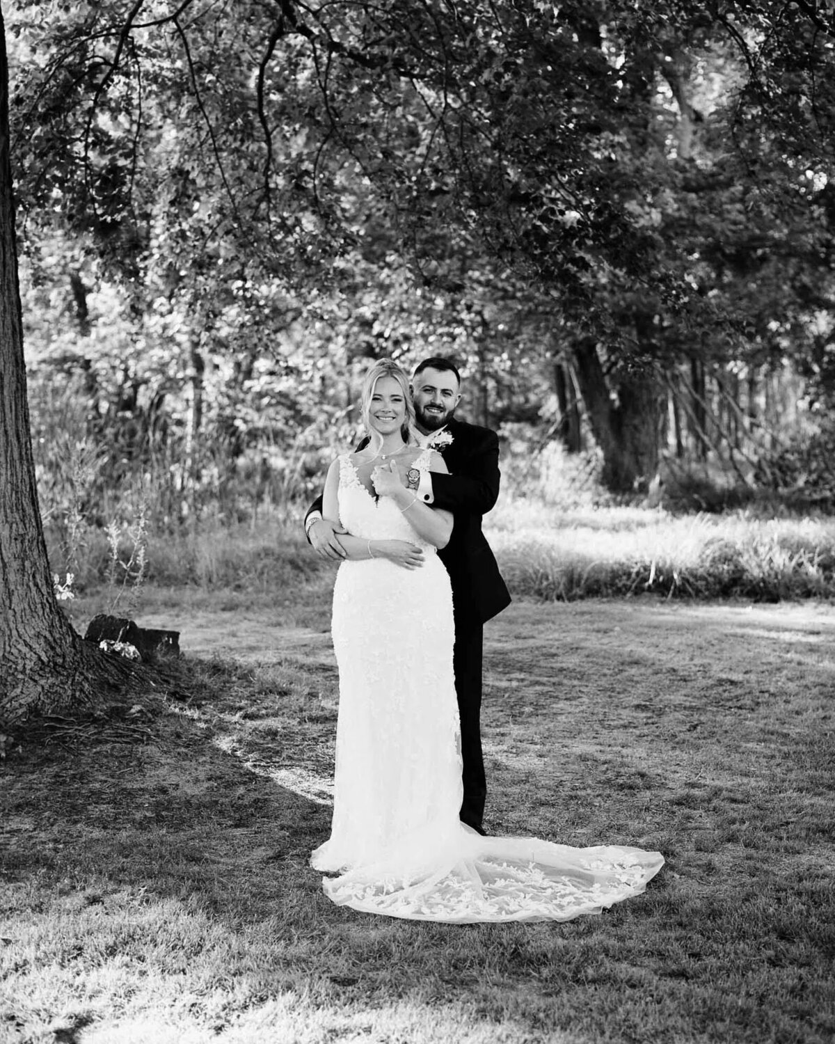 A bride in a white gown and a groom in a dark suit stand together outdoors, smiling and embracing under a large tree, with sunlight filtering through the leafy forest—captured beautifully by an NJ wedding photographer.