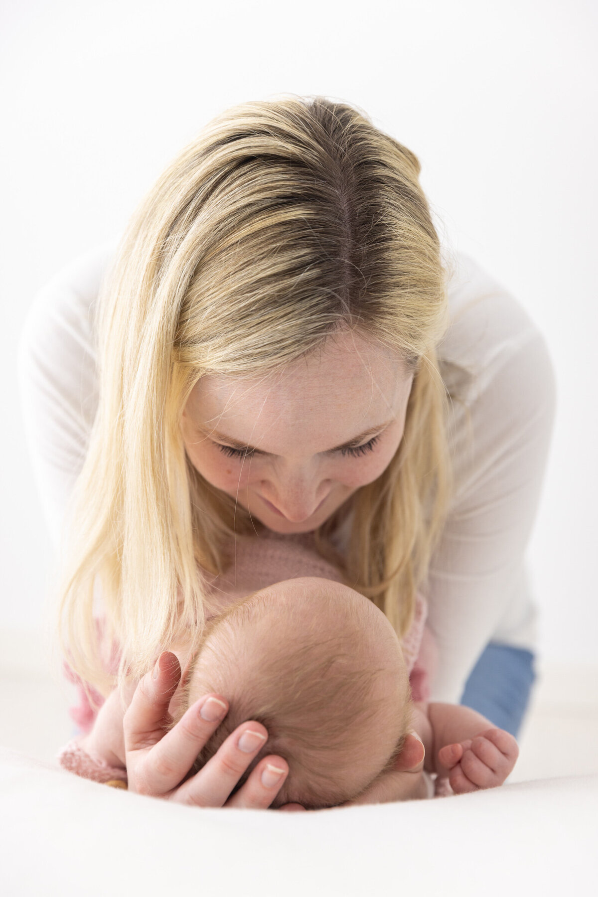 Mor ser på datteren sin. Hun bøyer seg ned over datteren som ligger på en stor hvit pute. Mors hender holder rundt hodet til datteren og man kan se hodet til babyen og mors hode/hår. Fotografert i Studio Landmark, Fyllingsdalen.