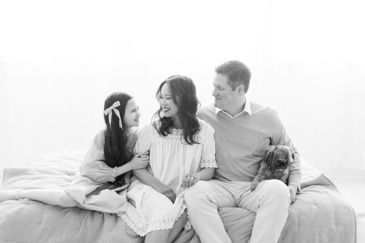 A Black and white portrait taken in a Dallas/Fort Worth portrait photography studio of a family of 3 with their dog as they are snuggled together on a bed for their new family photos.