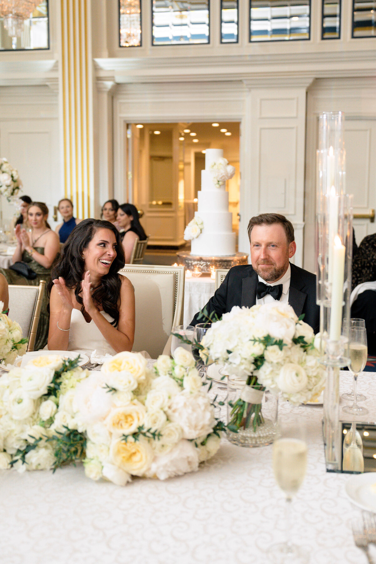 A bride and groom sit at a lavishly decorated table with white flowers and candles. The bride is smiling and clapping, while the groom looks content. A tiered white wedding cake is visible in the background. Guests are seated nearby.