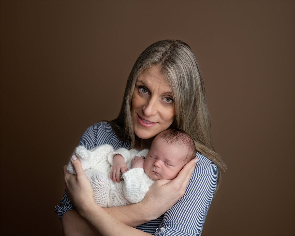 A studio portrait of a mother with soft blonde hair holding her newborn baby in a cozy embrace. Photographed at Meraki Photographic Creations in Paris, Ontario.