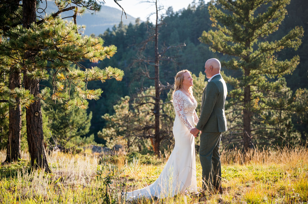 A bride and groom stand in a woodland Colorado setting, looking at each other hand-in-hand.