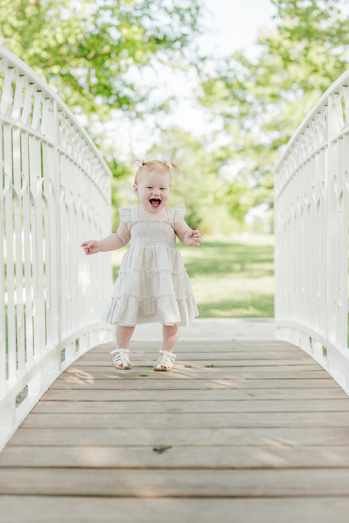 Little girl joyfully running across a white bridge in a light pink dress during a milestone photo session.