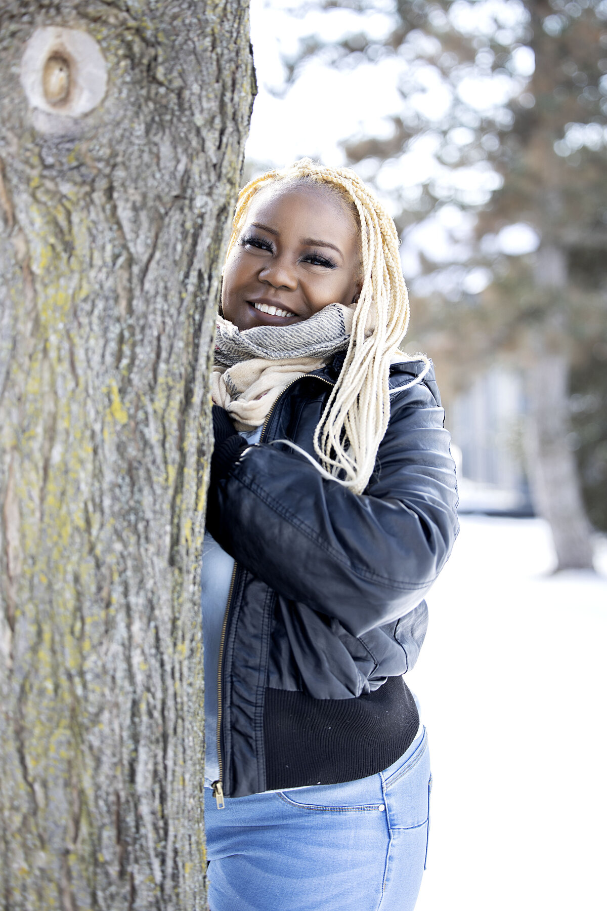 Woman in black leather jacket, blue jean top and jeans standing behind tree