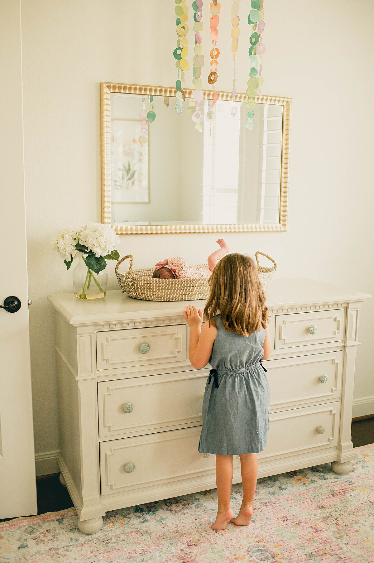 Little girl peeking over changing basket on dresser at new baby sister by Houston Newborn Photographer Mel B