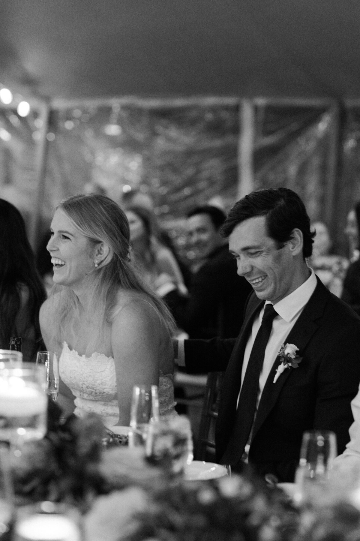 The bride and groom, seated at their wedding reception, share a laugh under a tent at Spring Lake Golf Club.
