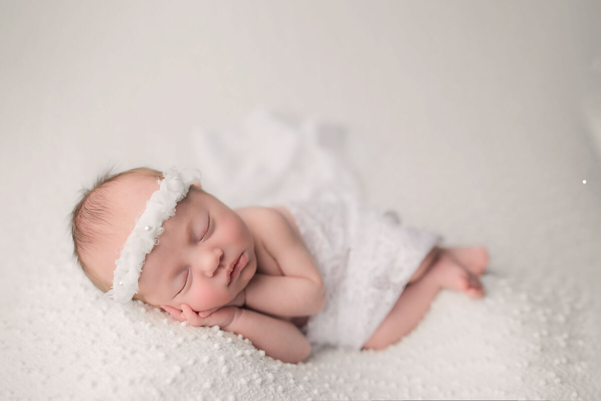 2 week old baby girl laying on side with hands together under cheek wearing white wrap and headband on white backdrop