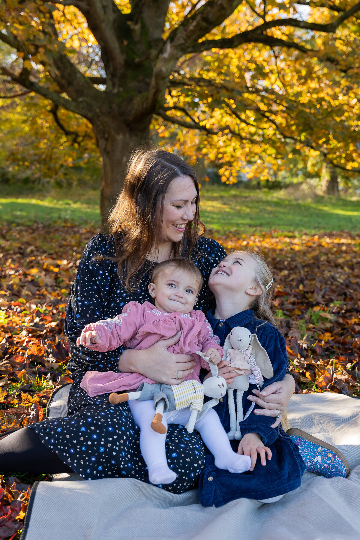 A mother embracing her two young daughters in a park with autumn leaves, smiling as the older child playfully kisses her cheek.