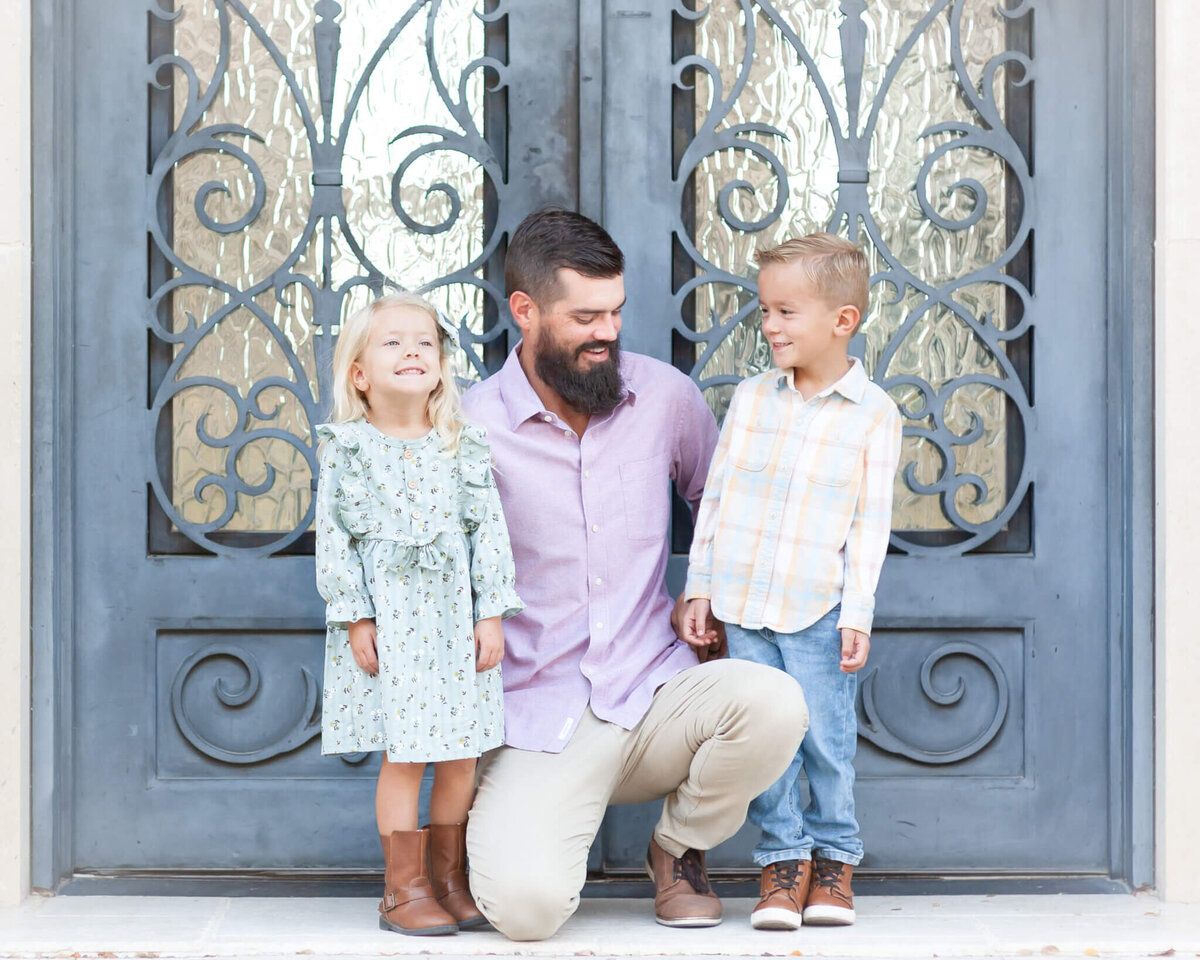 Father in  light purple crouched down with daughter and son in pastels next to a beautiful door in Adriatica
