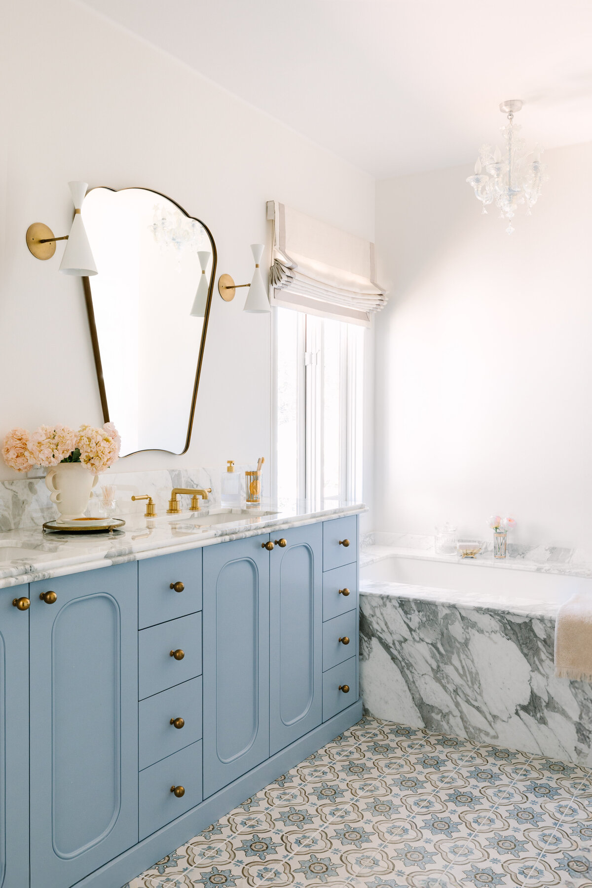 Primary bathroom with a custom vanity in Sherwin Williams Lakeside and rounded edges, Arabescato Corchia marble counters, Italian shield mirrors, Blueprint Lighting Monolith Wall Sconce, patterned floor tile, Murano chandelier, and a built in marble tub.