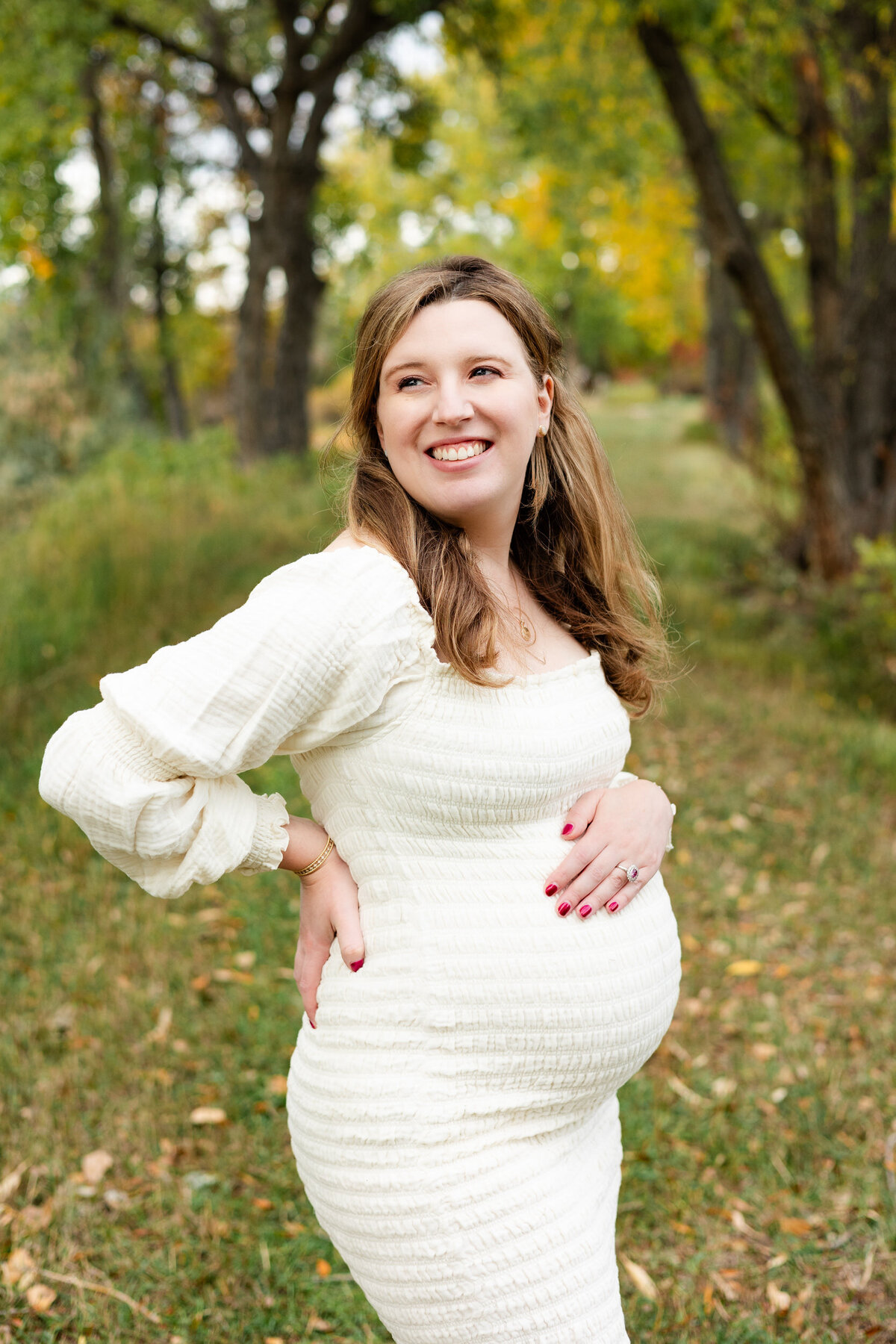 A pregnant woman has one hand on her back and one hand on her belly as she looks back over her shoulder and smiles.