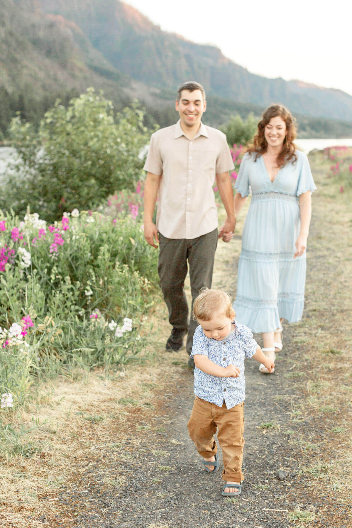 Toddler boy running in front of parents while Mom and Dad hold hands and walk behind him smiling. They are walking next to a patch of pink wildflowers with the river off to the side and the high cliffs of the Columbia River Gorge behind them.