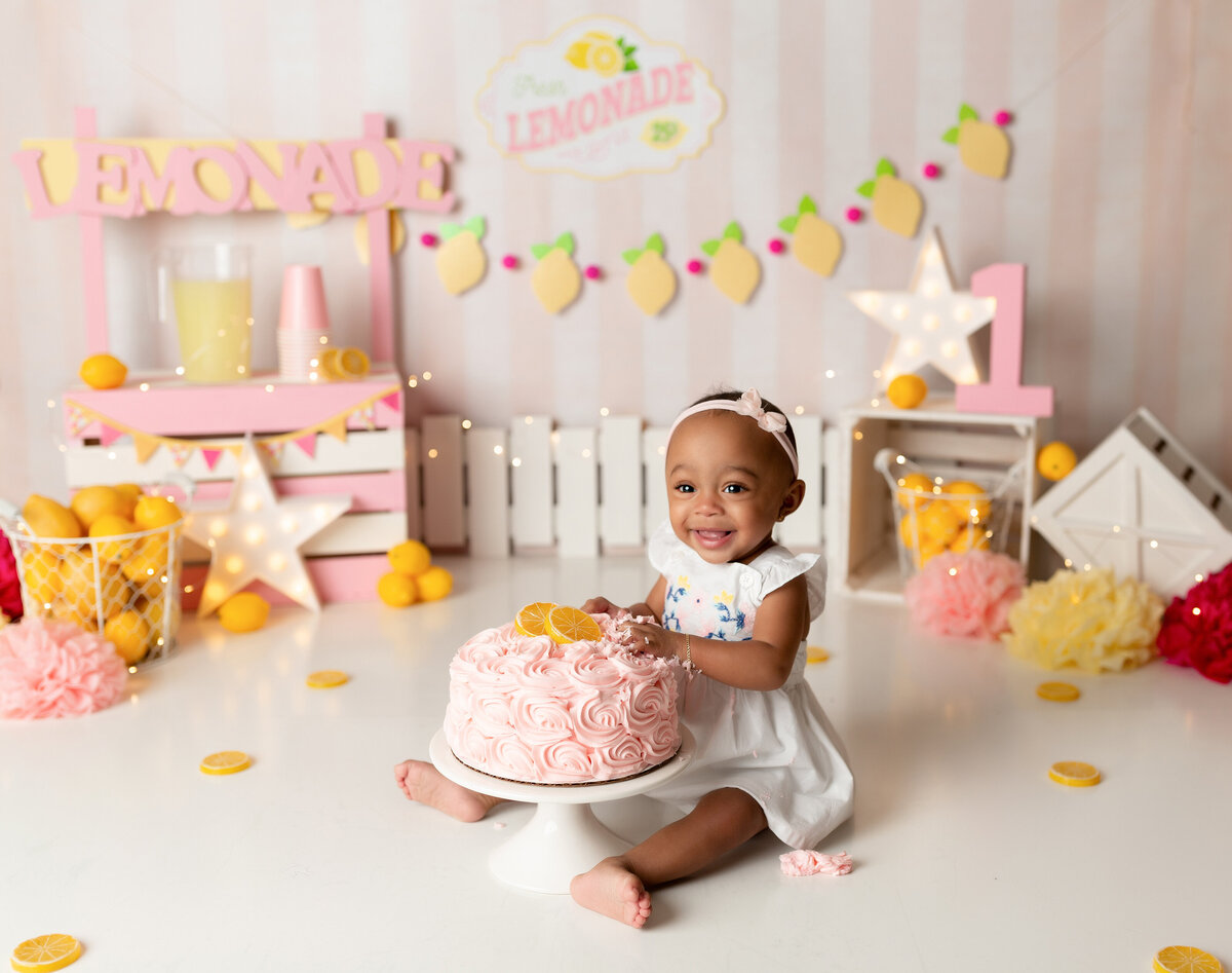 Lemonade stand themed cake smash in West Palm Beach, and Boynton Beach, FL photography studio. Baby girl has her hands in a pink rosette cake topped with lemons. She is smiling at the camera. In the background, there is a pink and white lemonade stand, lemons, and flowers.