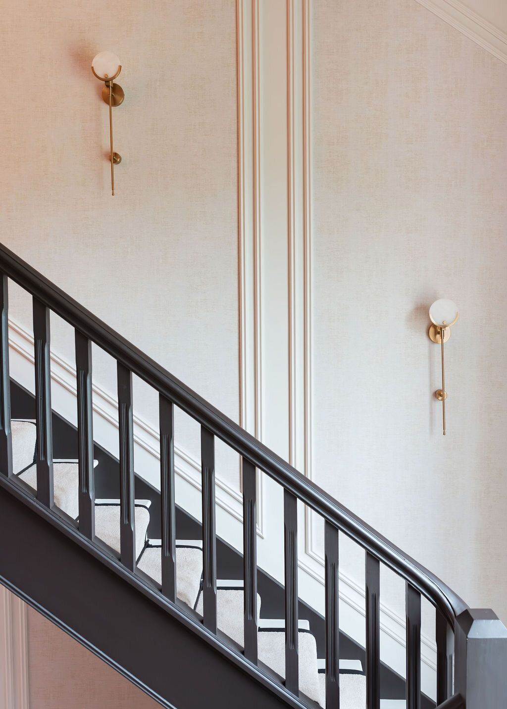 A minimalist interior features a staircase with dark wooden balusters and light-colored stairs. The walls are decorated with vertical molding and two spherical wall sconces with brass accents, creating a clean and elegant look.