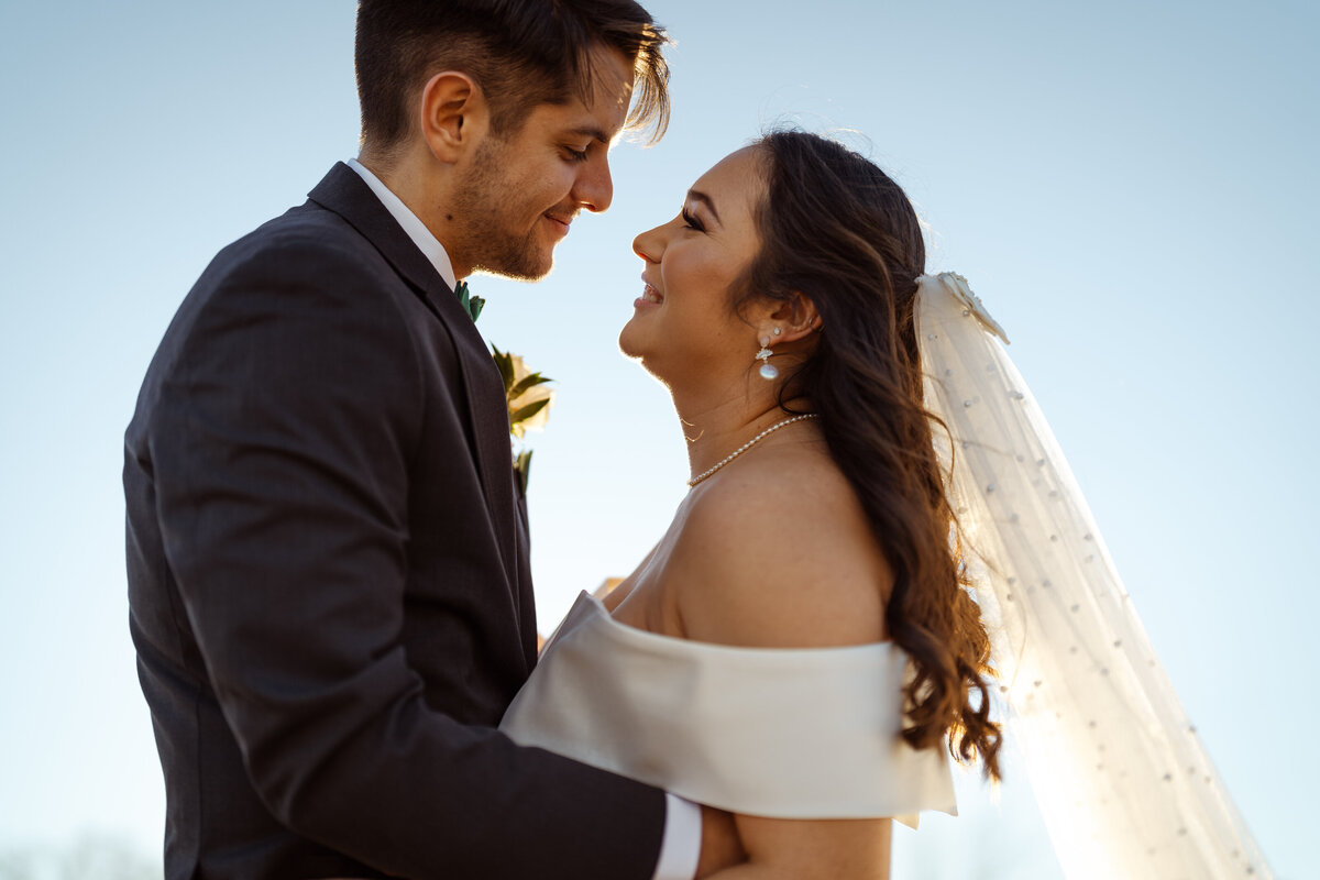 Close-up photo of a newly married couple holding each other and staring lovingly into each other's eyes as they smile during a winter sunset at D'Vine Grace Vineyards in DFW, Texas. The image captures their deep connection and the romantic ambiance of the moment against the sky