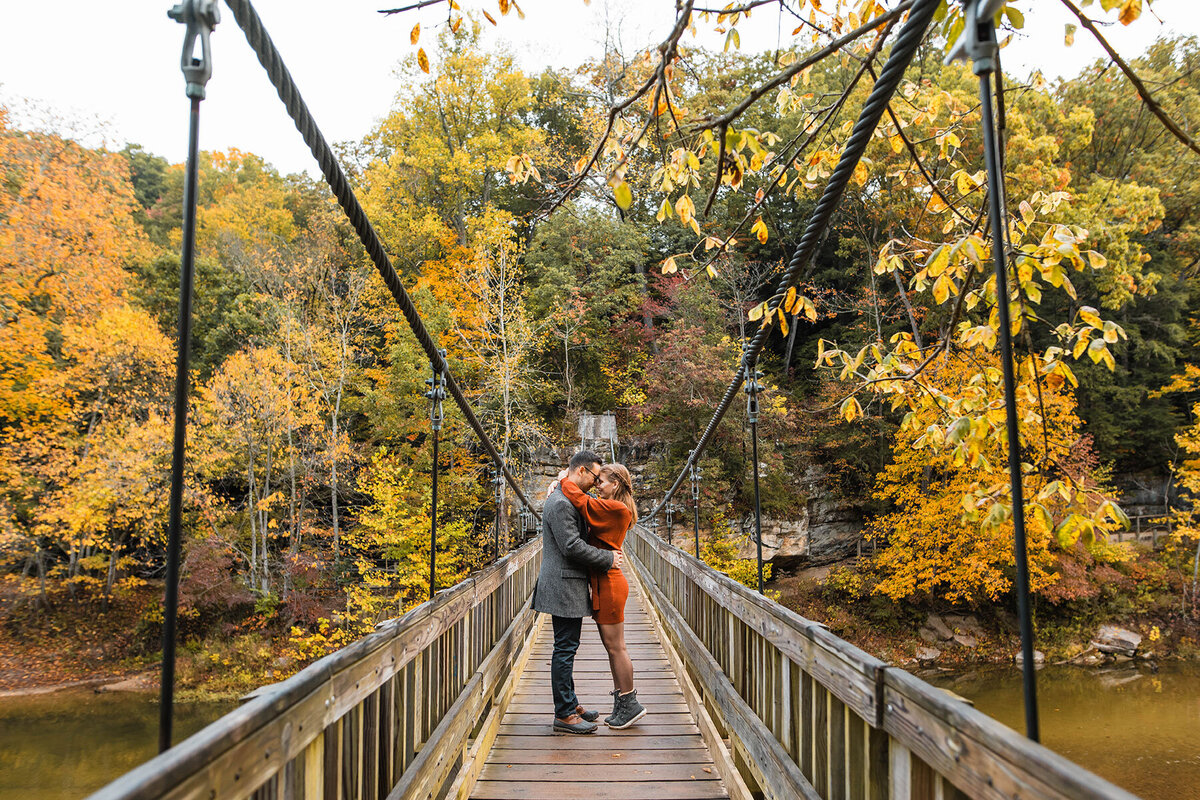 Couple rests heads together while standing in the middle of a suspension bridge  surrounded by fall leaves.