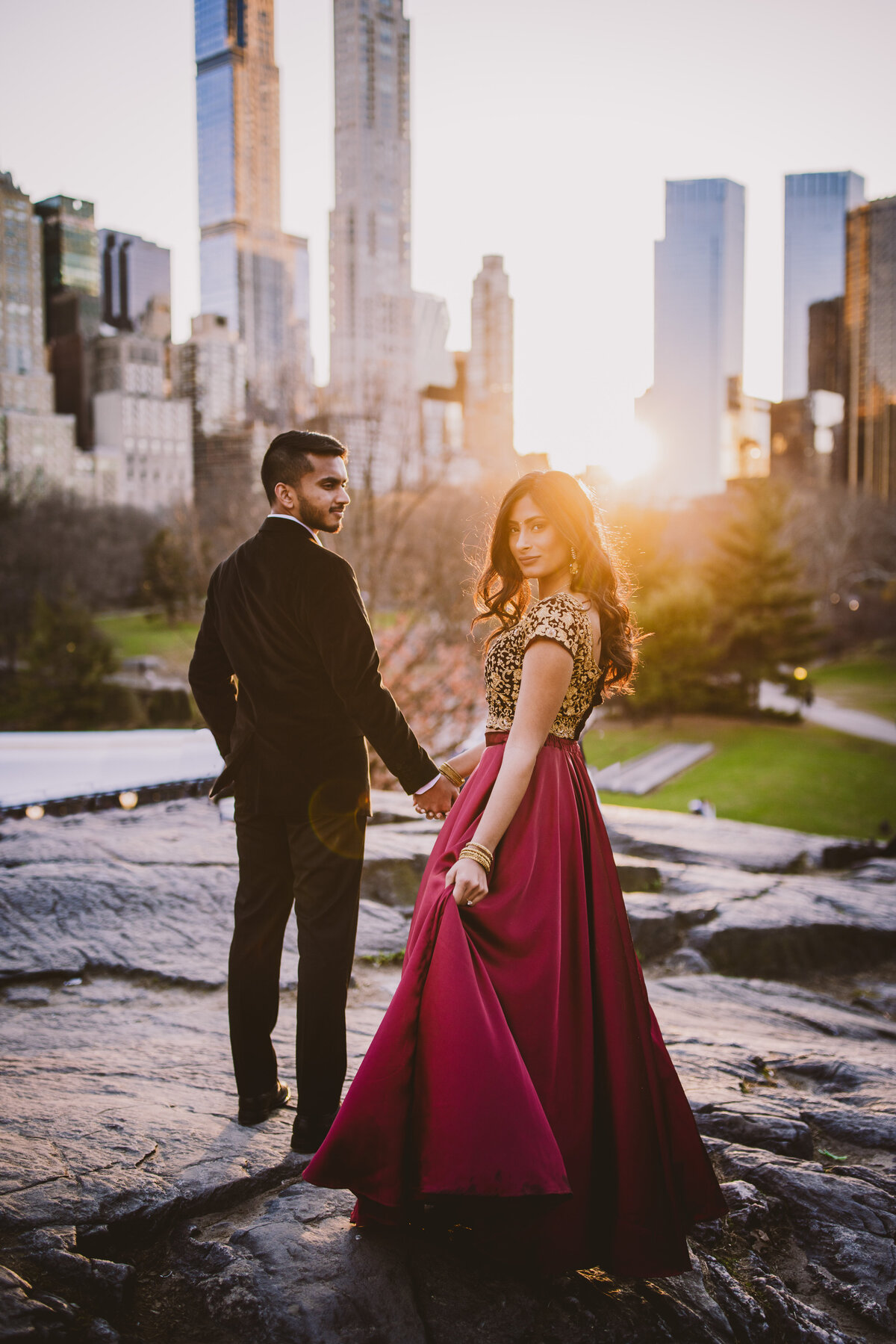 An engaged couple in indian clothing and a suit look back at the camera in front of the nyc skyline on a rock at central park