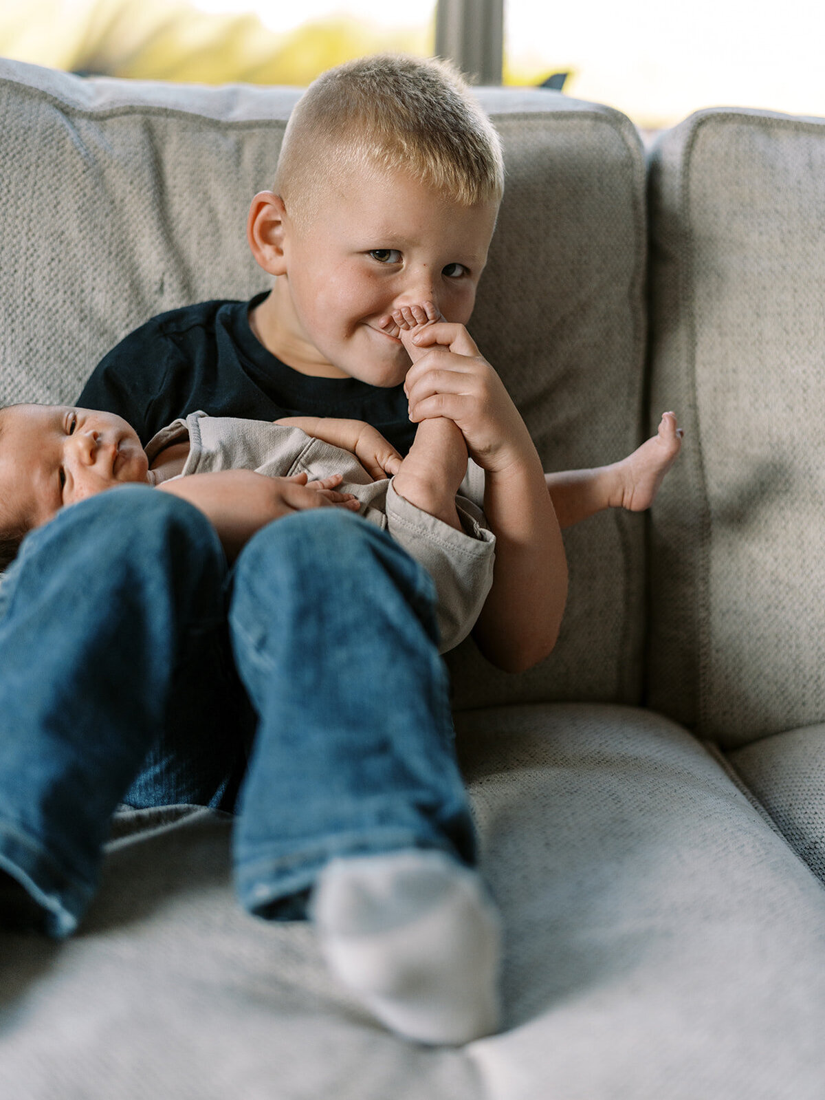 A young boy holding a baby sits on a couch at home and smiles at the camera.