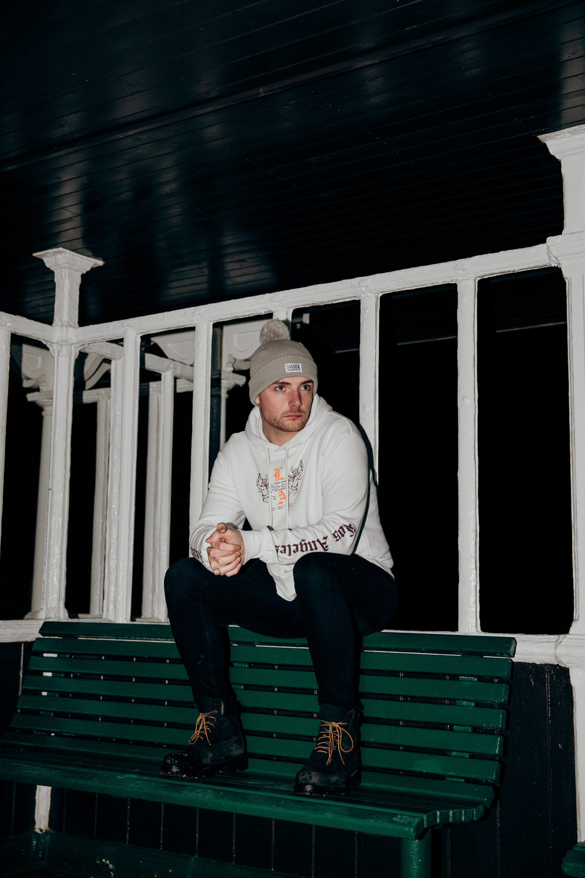 Connor sat on top of a bench under a shelter on Brighton Beach, shot at night with flash.