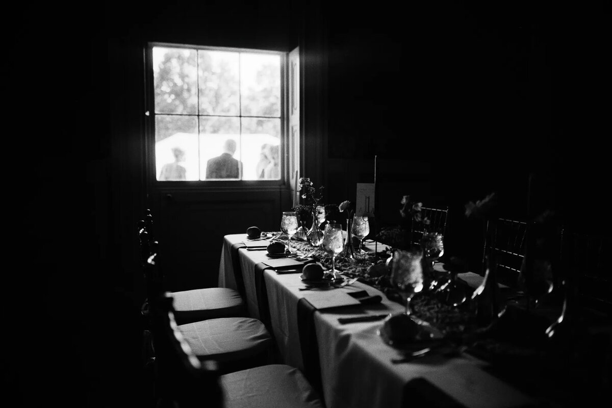 A dimly lit dining table set with glassware and plates sits in the foreground, beautifully captured by a film photographer NJ, while the silhouettes of people are visible outside a bright window in the background.