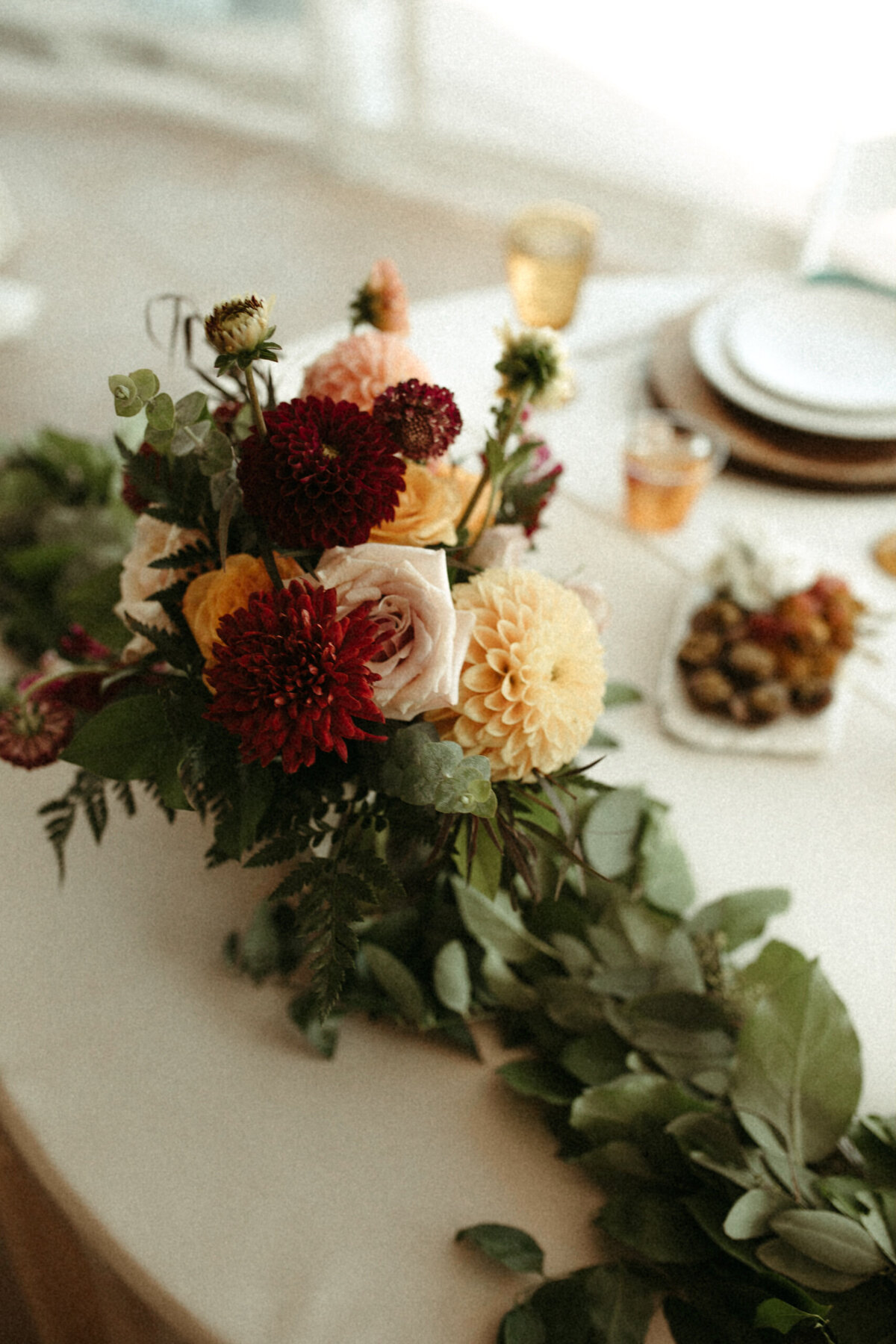 Burgundy, yellow, and blush flowers on a sweetheart table with a greenery runner at a wedding reception.