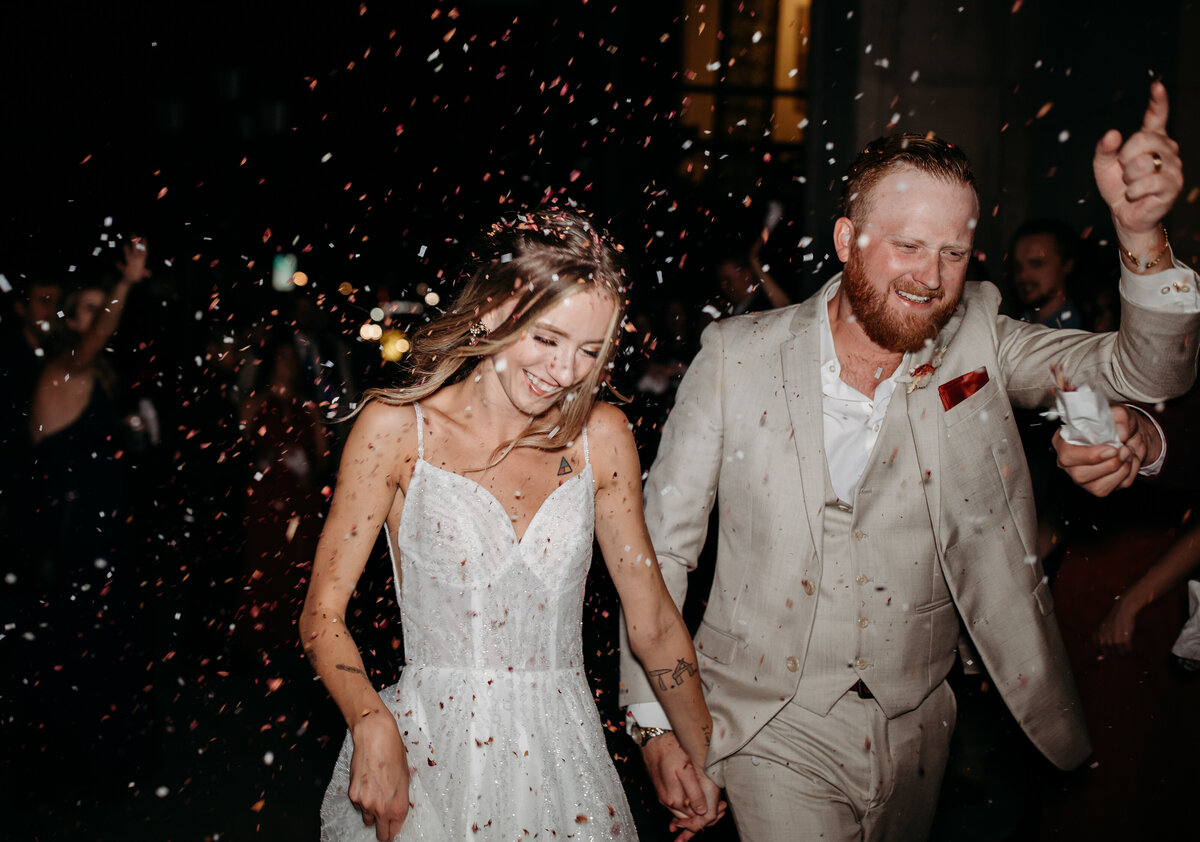 Newlywed couple joyfully walking through a shower of confetti, the groom in a beige suit and the bride in a sparkling gown