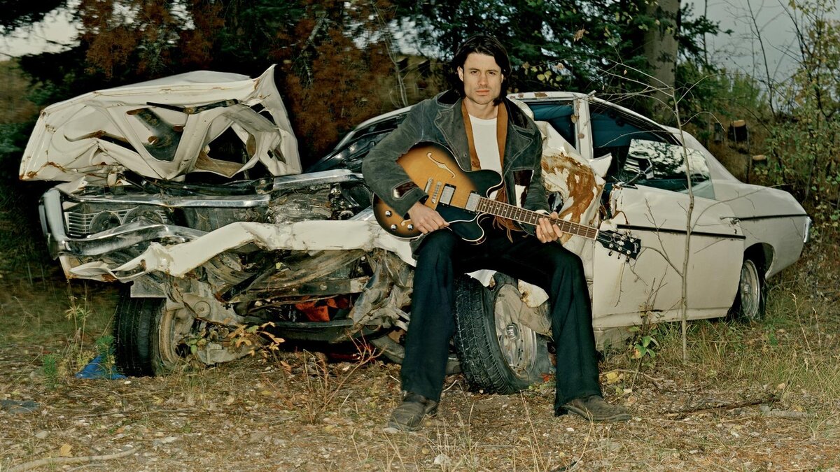 Musician portrait Dustin Bentall sitting on fender of wrecked car guitar in lap Roadshow Gallery