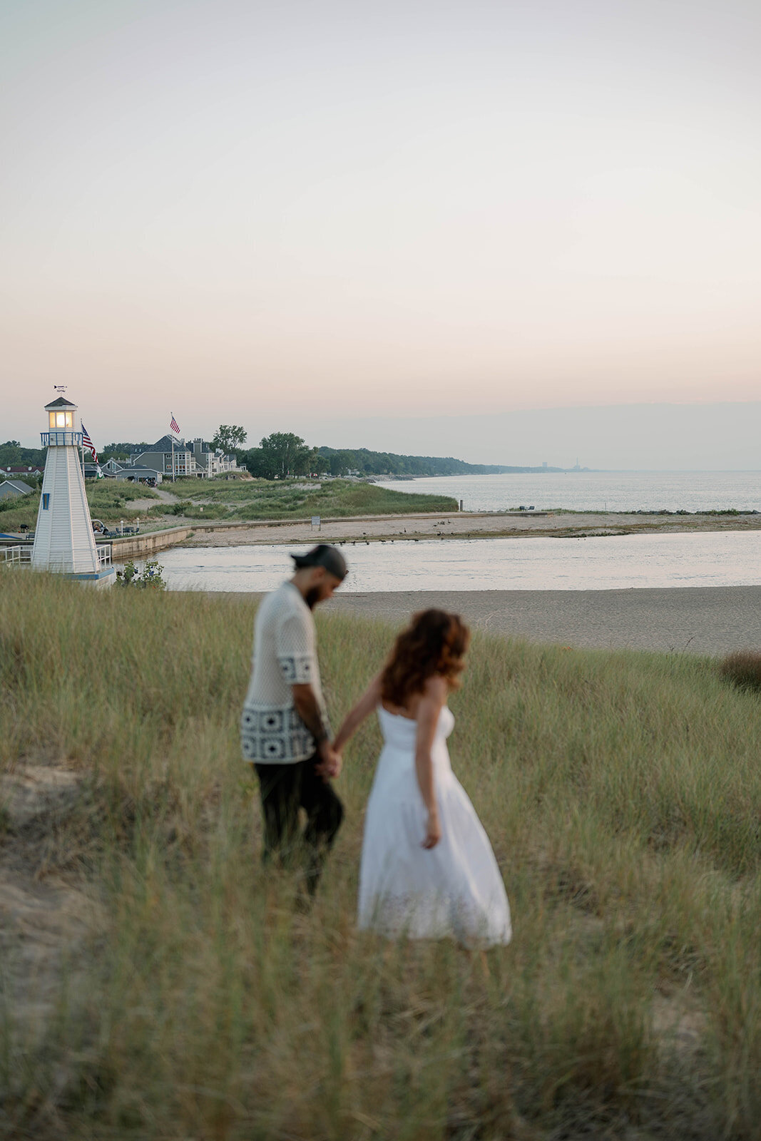 Engaged couple walking through the New Buffalo dunes with the lighthouse in the background during sunset engagement photos