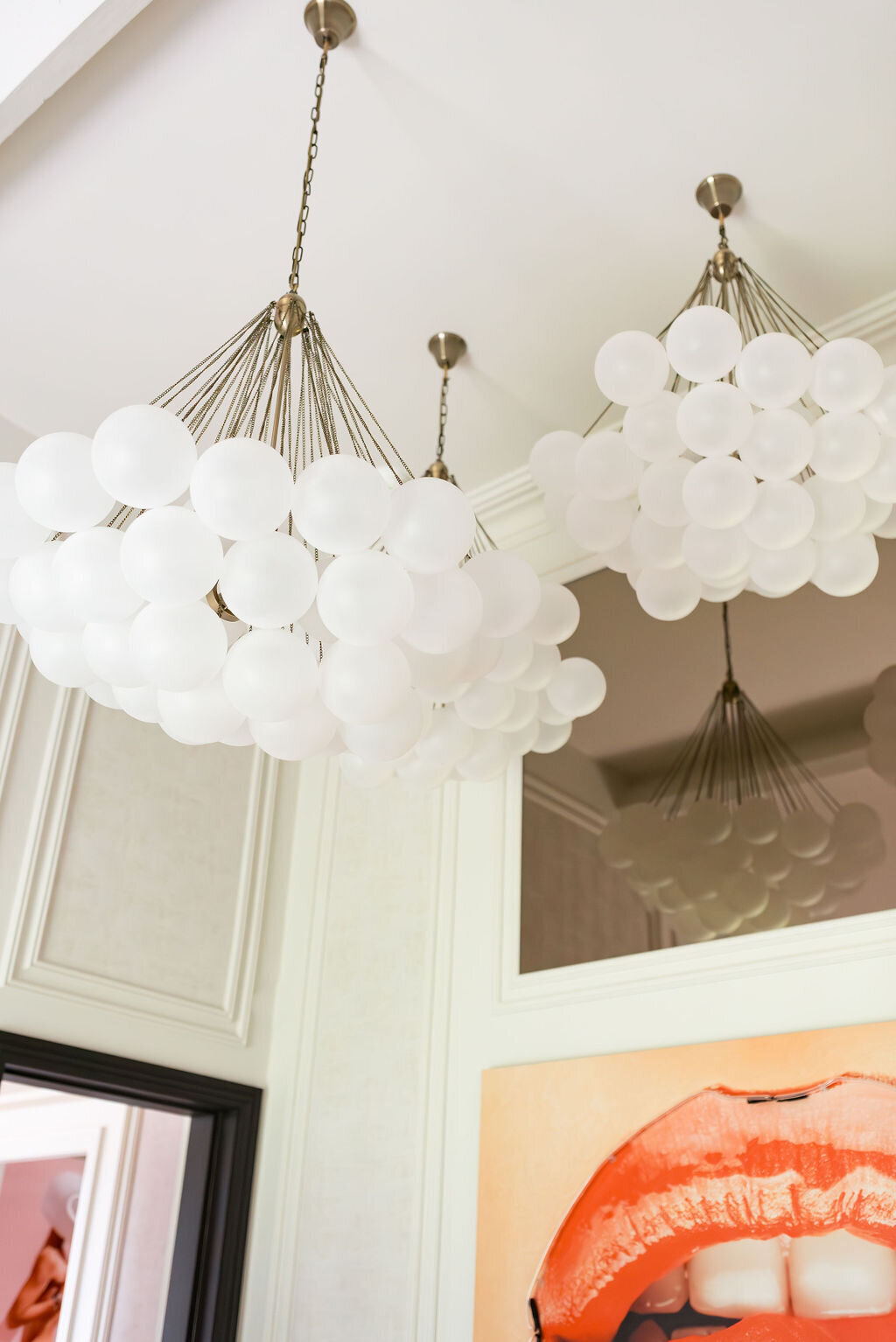 Three modern chandeliers with frosted white glass globes hang from the ceiling of a room with light-colored walls. Below them is a framed artwork featuring a close-up of vividly colored lips, adding a splash of color to the space.