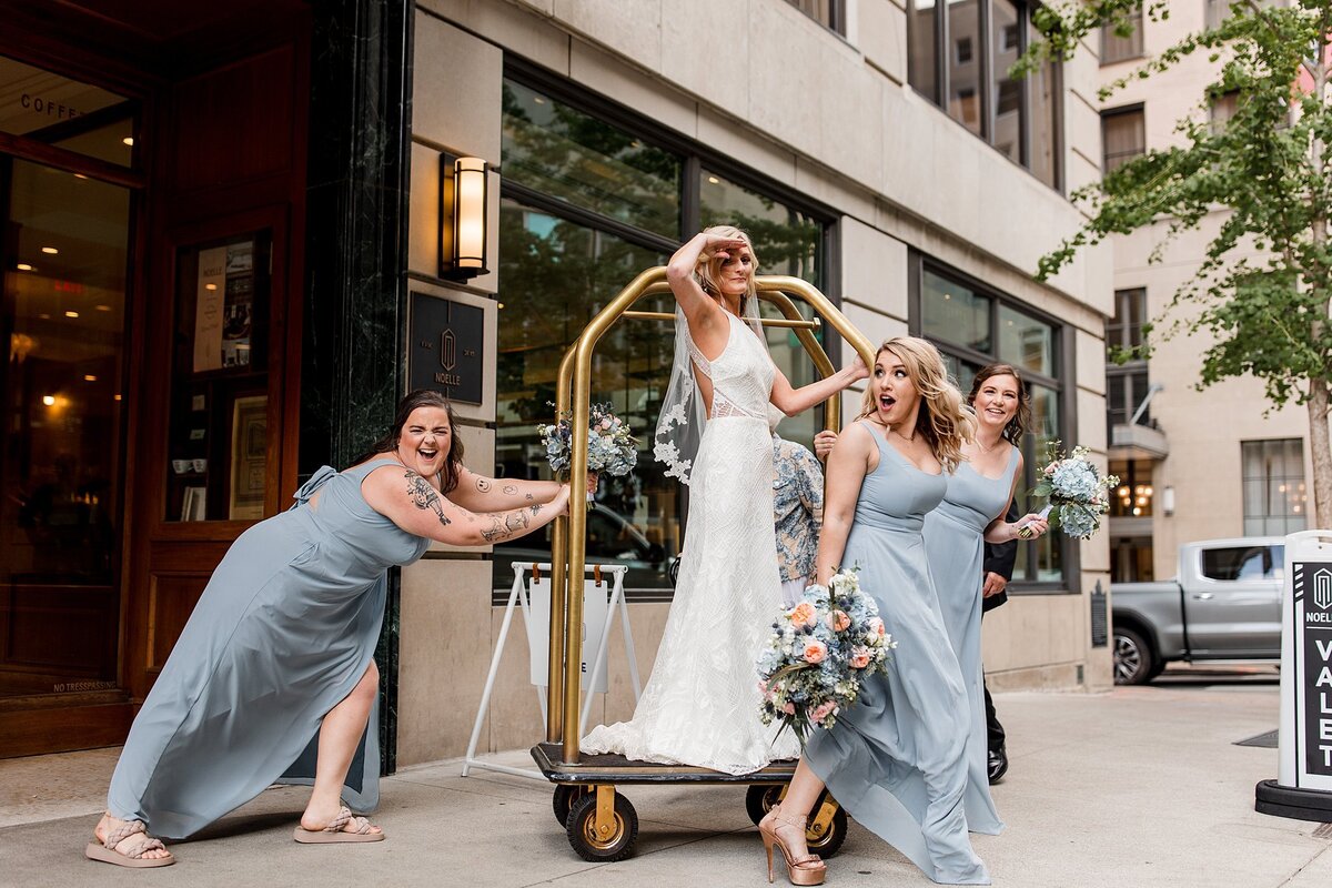 Bridesmaids in light blue dresses holding bouquets of pink and white roses with greenery push the bride wearing a halter top wedding dress and veil on a gold luggage cart in front of the Noelle Hotel Nashville.