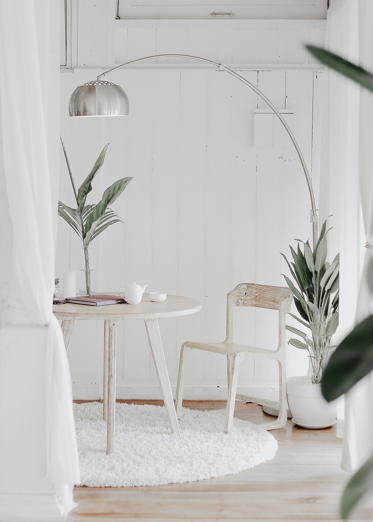 A white washed wooden wall is pictured in a corner with boho distressed table and chair with plants and a metal floor lamp.