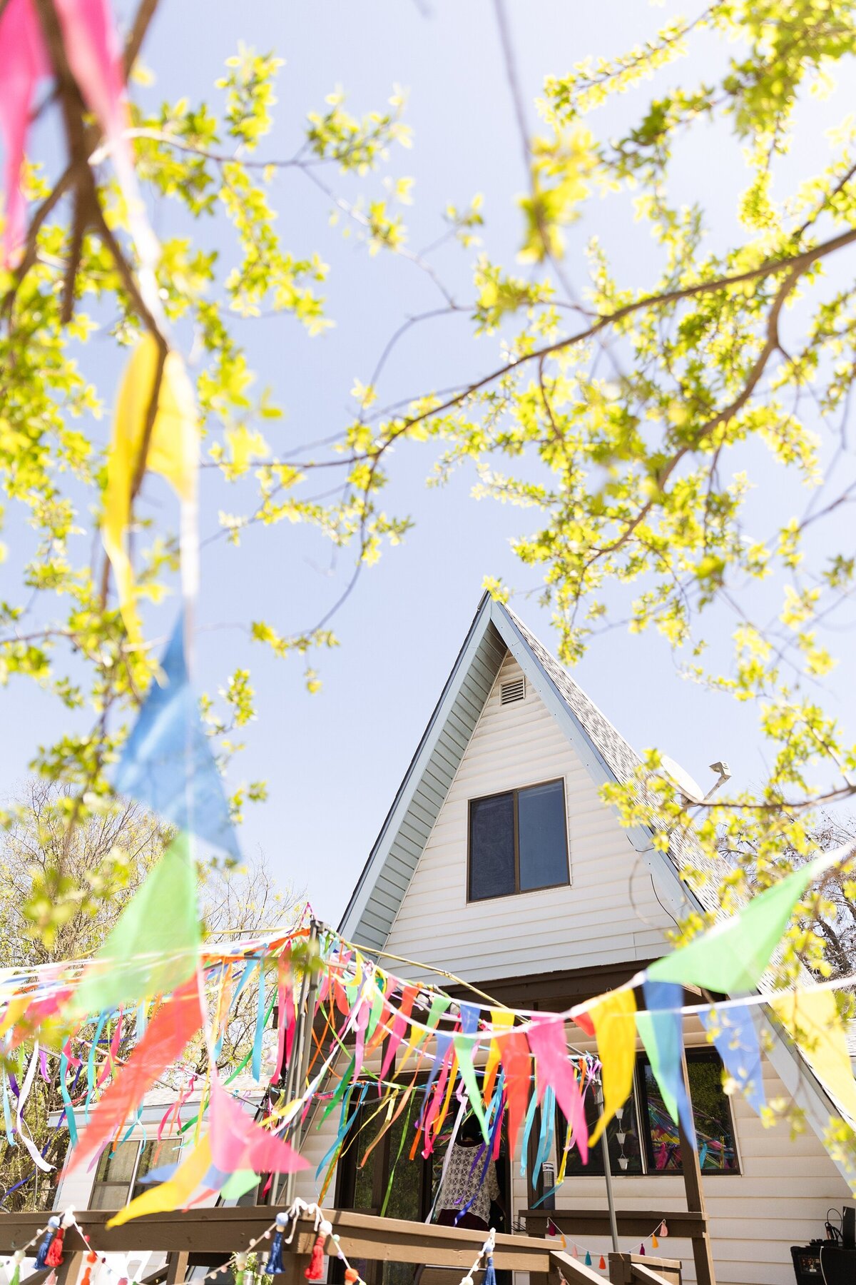 A detail shot of a backyard wedding reception in Dallas Fort Worth, Texas. Many lines of colorful flags adorn the backyard porch which is surrounded by the branches of many trees and backed by the triangular house in the background.