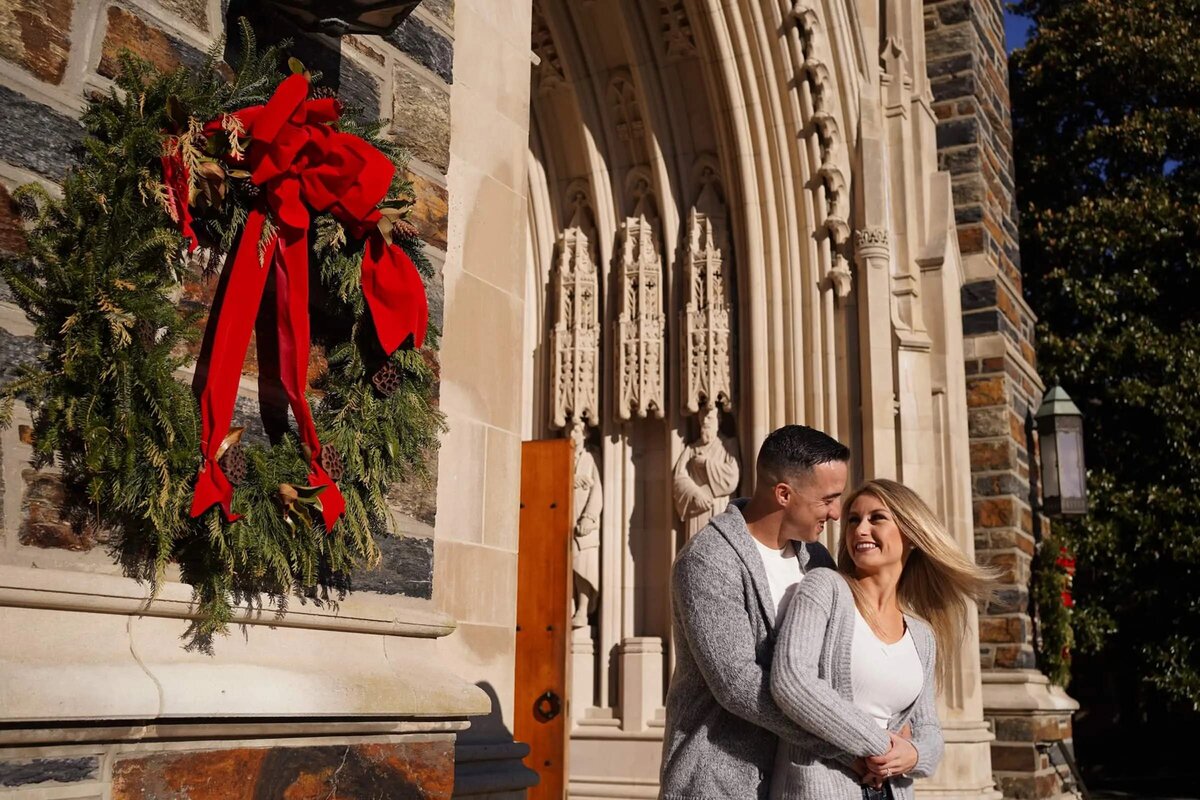 A couple stands close together in front of an ornate building adorned with a red Christmas wreath