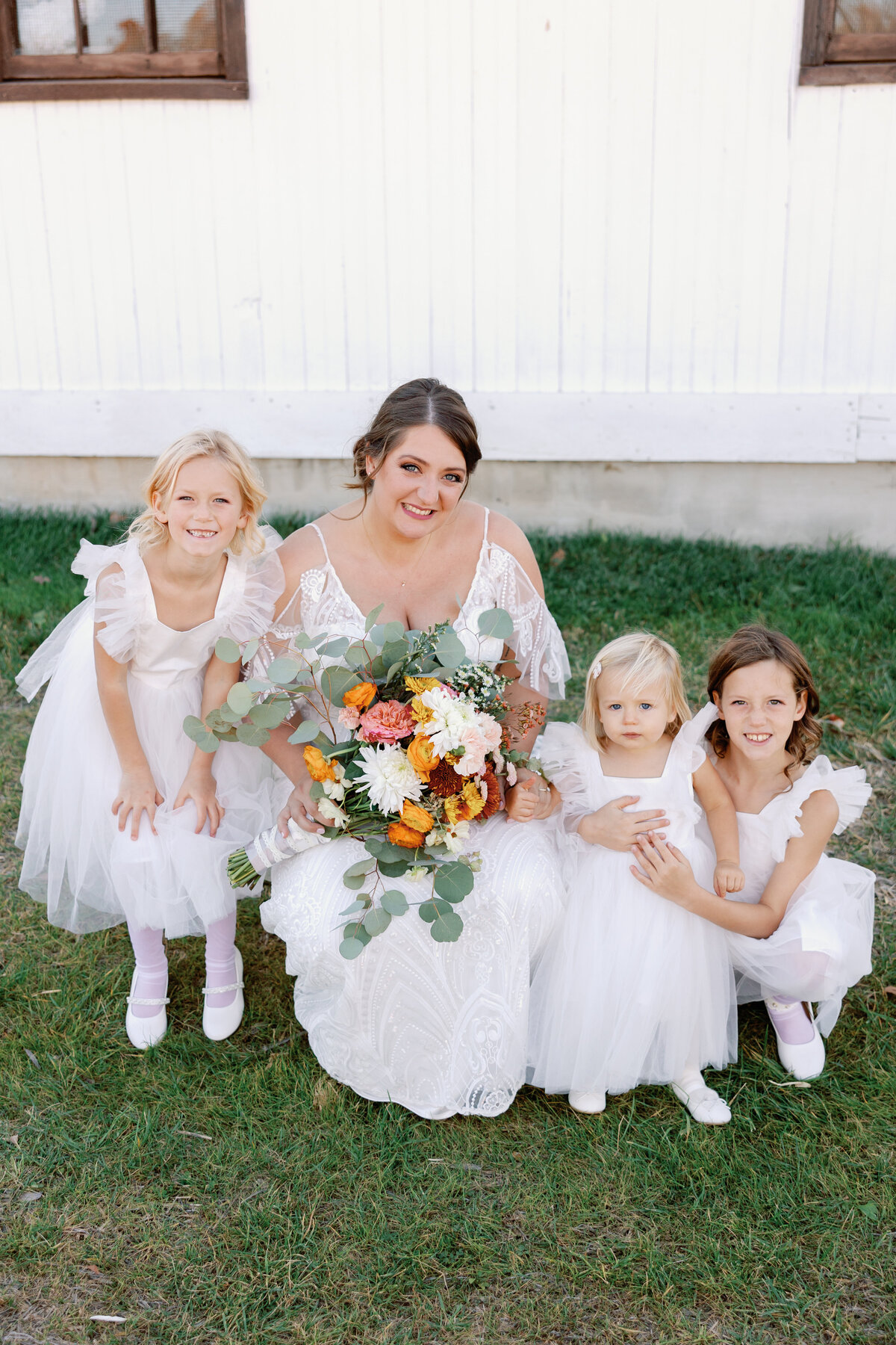 Bride in lace gown sitting with three flower girls in white tulle dresses, holding colorful bouquets.