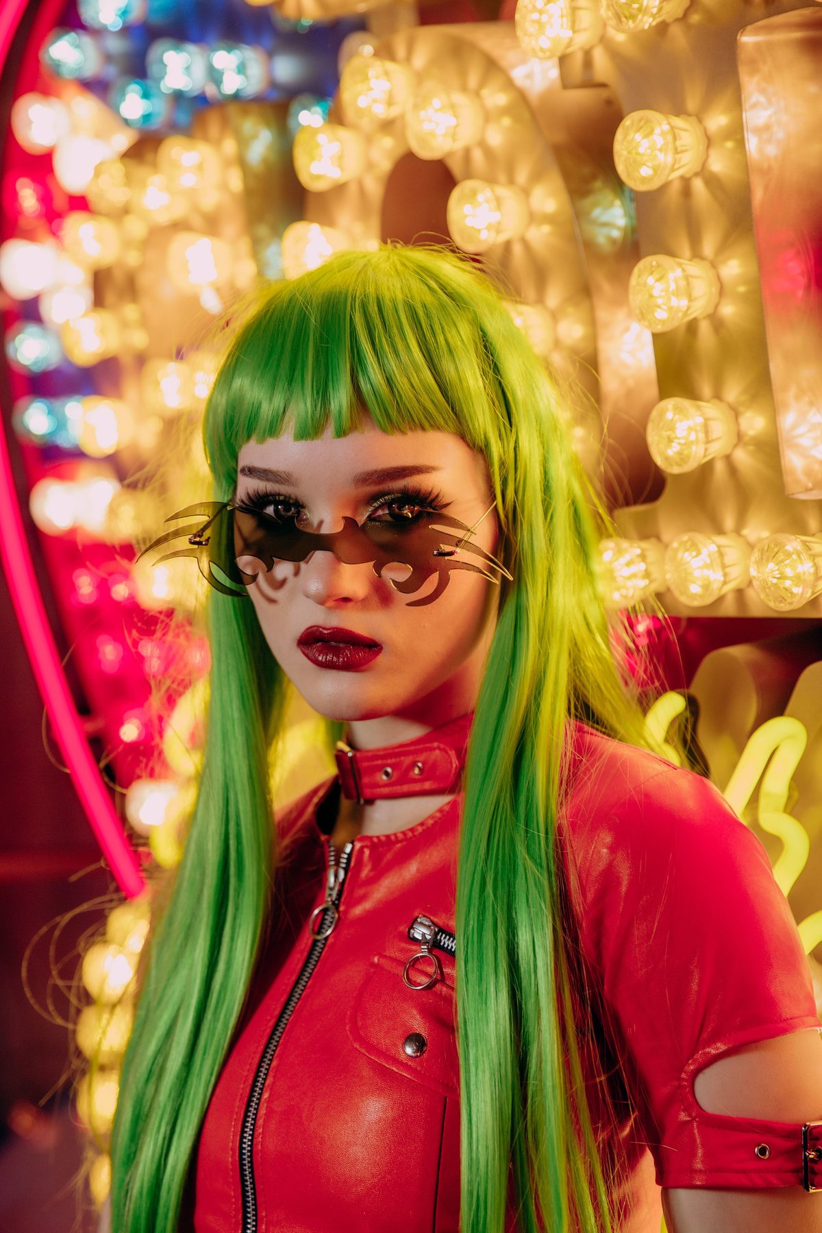 Saida leaning against an old school light-bulb sign, wearing  a red leather dress, a green wig and unusual sunglasses