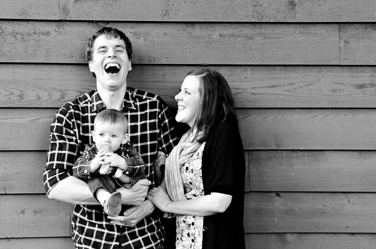 The black-and-white image shows three people standing in front of a wooden plank wall. On the left, a man wearing a checkered shirt is holding a baby. The baby is wearing a checkered shirt, jeans, and shoes and is holding a shoe to their mouth. On the right, a woman with long hair, wearing a patterned dress and a dark cardigan, is looking at the man and smiling. The man is laughing with his mouth wide open.