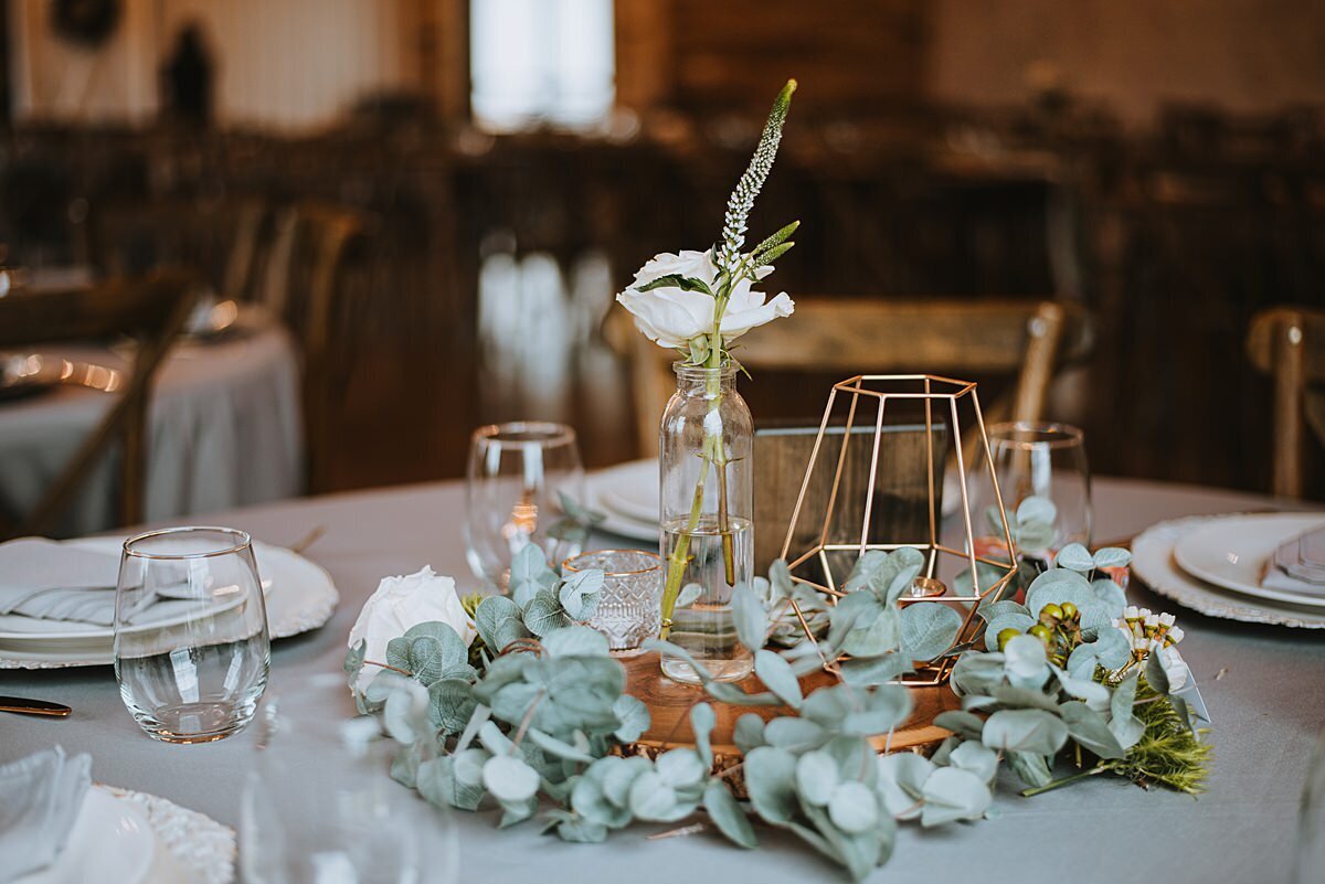 A wood slice encircled by eucalyptus and topped with a clear glass bud vase with veronica and a white  ranunculus is set next to a brass wire geometric candle holder in the center of a table set with ivory chargers, white plates and stemless wine glasses at Steel Magnolia Barn.