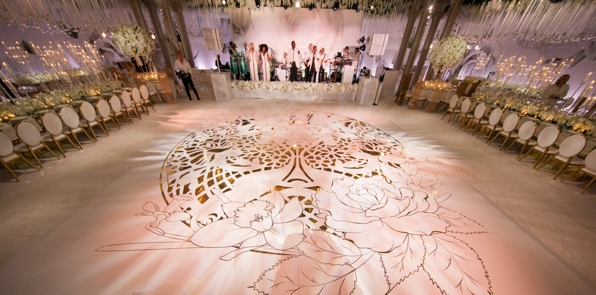 The photo was captured at The New York Public Library in New York City by photographer Christian Oth, showcasing a grand wedding venue with elegant white and gold decor. The focal point of the scene is a large, intricate floral pattern on the dance floor, adding a touch of sophistication to the setting. A live band can be seen performing on a beautifully decorated stage, while flowers serve as a charming backdrop. The elegant setup includes round tables adorned with white chairs on both sides, creating a stylish and inviting atmosphere for the guests. Additionally, East West Films provided videography services to capture the memorable moments of the event.