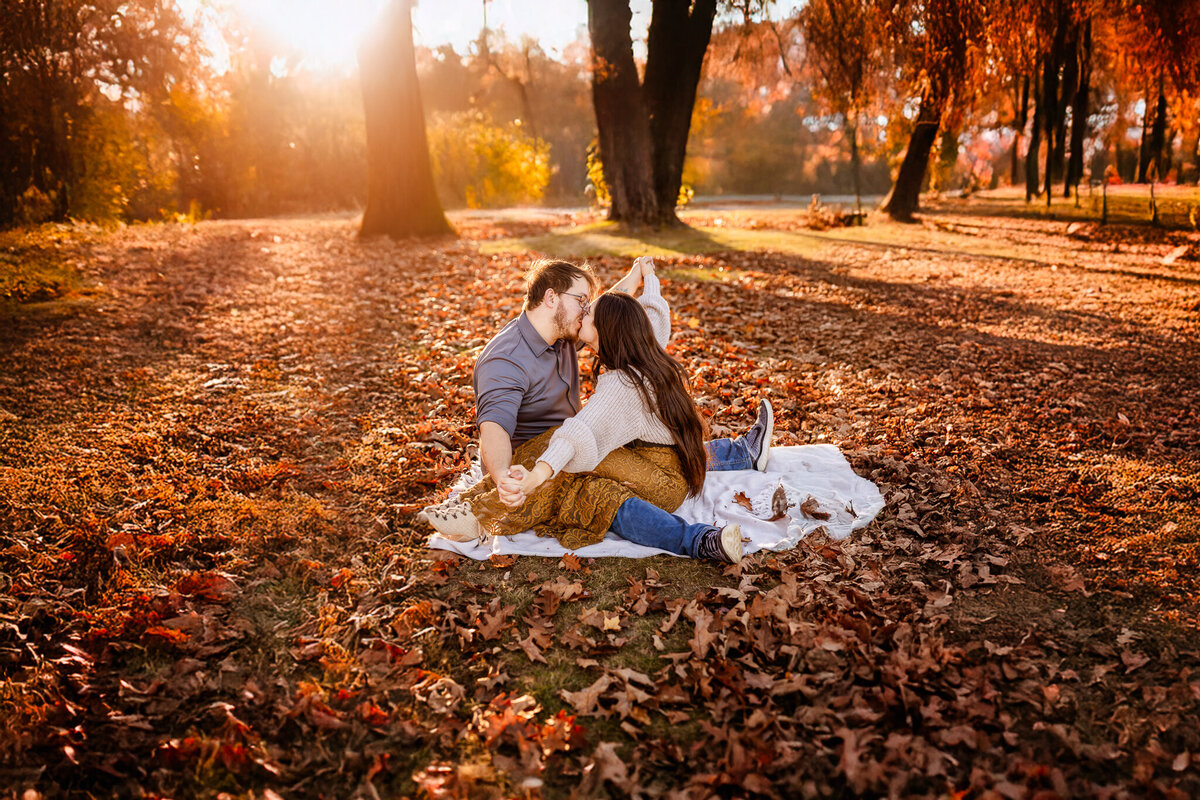 Couple kissing and sitting on blanket surrounded by vibrant fall leaves at sunset engagement photo session