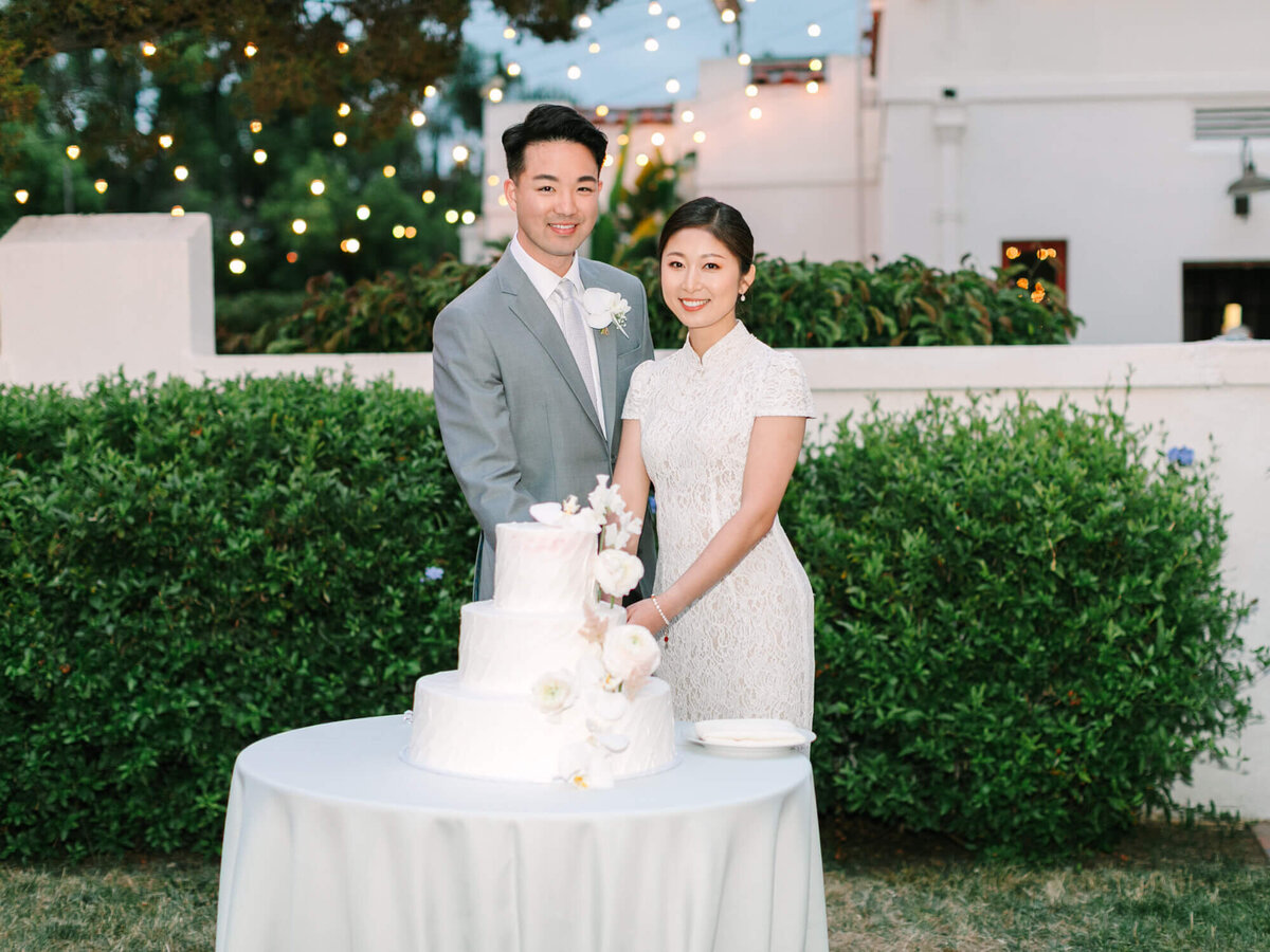 A smiling bride and groom stand beside a three-tiered white wedding cake adorned with flowers. They are in a garden setting with string lights.