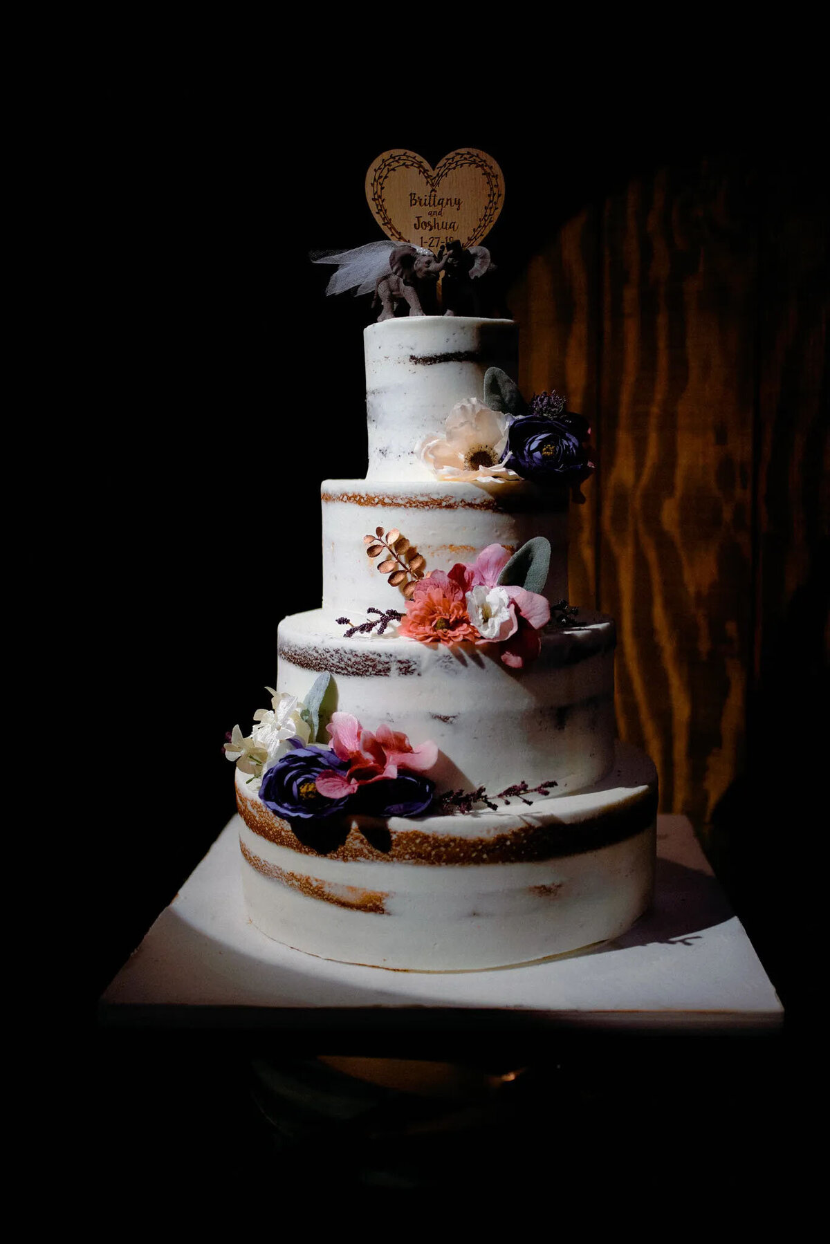 A four-tiered semi-naked wedding cake decorated with pink, white, and purple flowers, topped with a wooden heart-shaped “Bridging Healing” sign, beautifully captured by an NJ wedding photographer against a dark background.