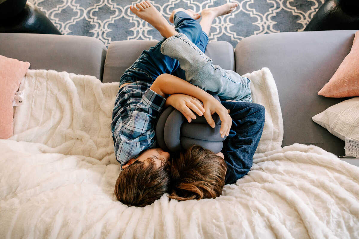 Two boys are tangled together on their Brookline living room couch: all we see is the tops of their heads and a tangle of feet and legs from above.