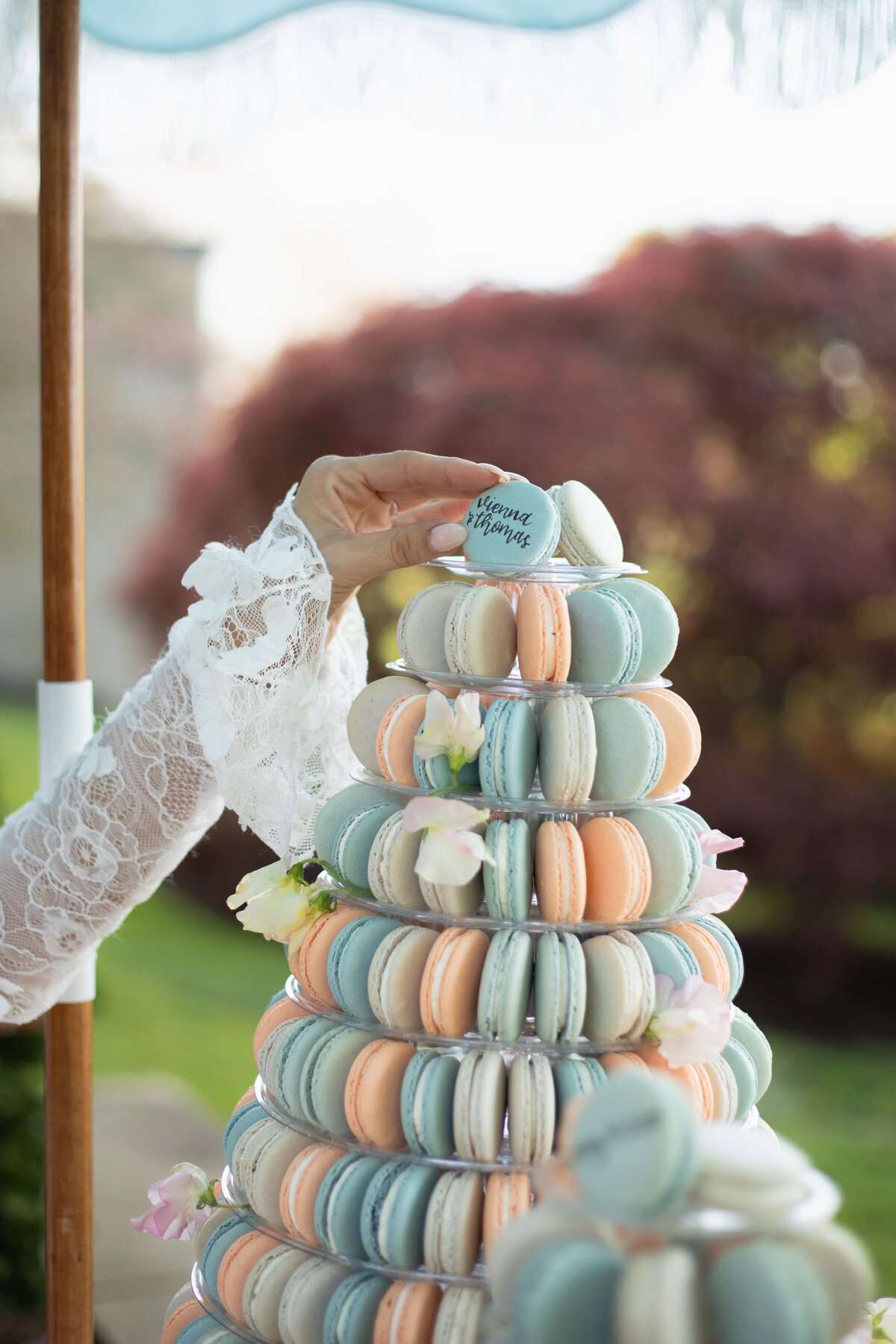 Wedding Photographers in RI | Kelsey Sheehan Photography Timeless Rhode Island Weddings | A hand in a lace sleeve places a blue macaron atop a tiered tower of pastel macarons, adorned with white flowers, outdoors in soft-focus greenery.