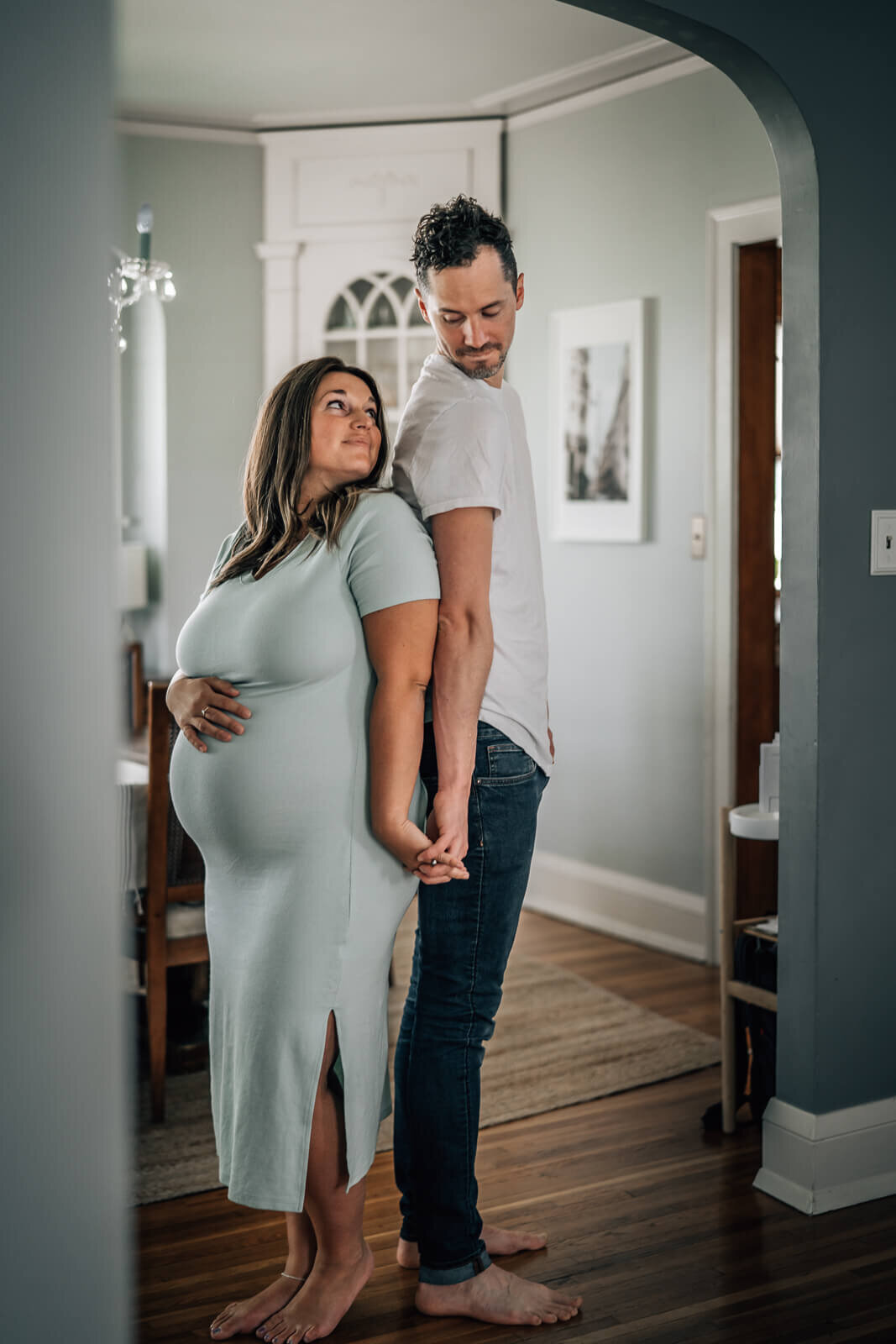 A pregnant mama stands back to back, holding hands and gazing up and back at her husband, frames by a beautiful architectural arch in their home.